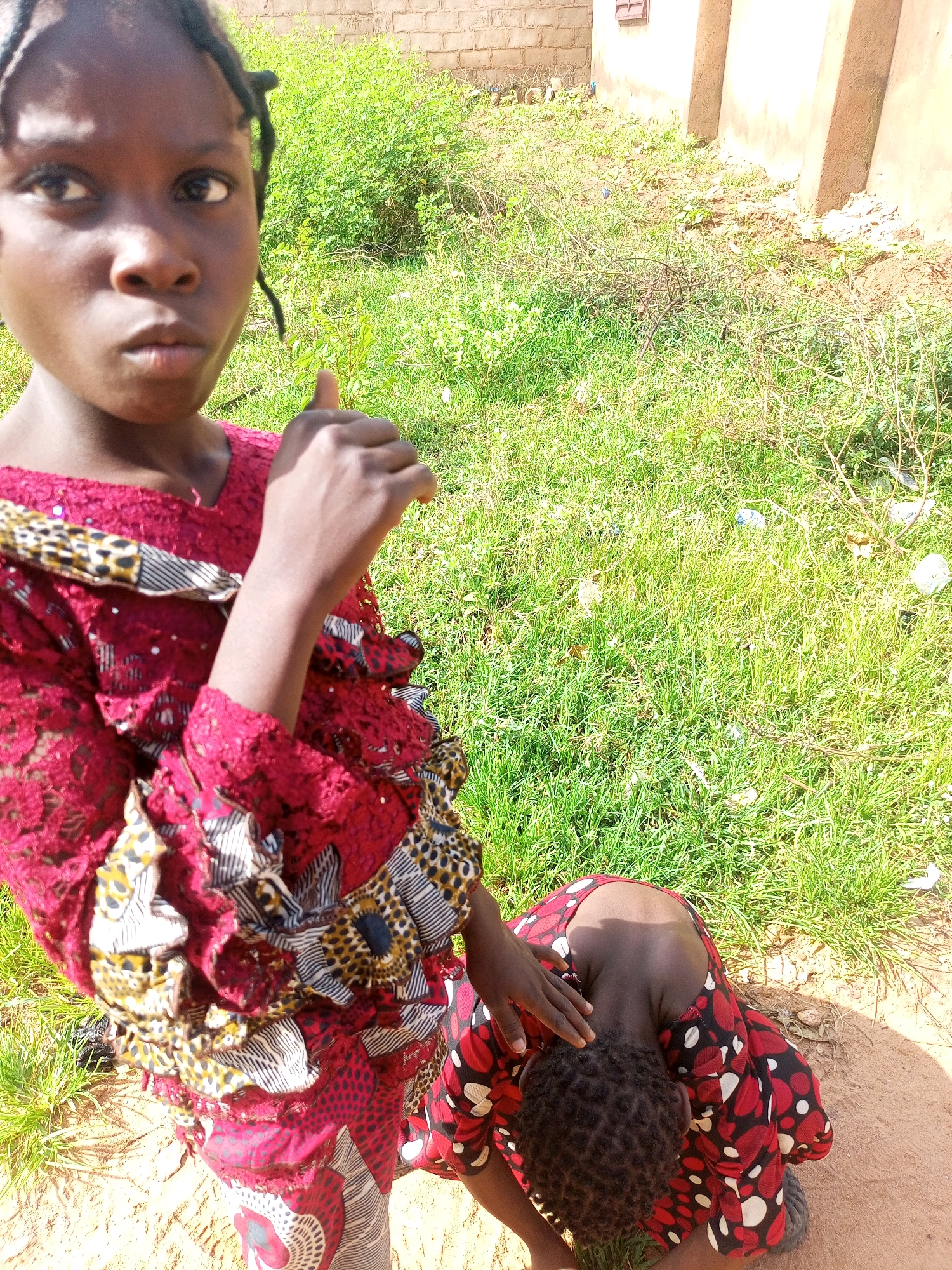 In the city of Tempure, Nigeria, two young individuals are present in an outdoor environment. One person, appearing to be a young girl, stands and looks directly at the camera, her face framed by braided hair. She is wearing a red and black patterned dress with intricate lace detailing and leopard print accents. Her right hand is raised, with her thumb extended in a thumbs-up gesture.
Kneeling in front of her is another individual, also appearing to be a young girl, whose head is bowed. She is wearing a red and black polka-dotted dress. The standing girl's left hand is resting on the kneeling girl's head, a gesture that could imply comfort or guidance.
The background consists of dry, sandy ground scattered with patches of green grass and some dry twigs. A brick wall and a light-colored structure are visible in the distance, suggesting a residential or semi-urban setting. The overall lighting indicates it is daytime, with shadows cast on the ground. The presence of some litter, such as plastic bottles, is noted on the grass.