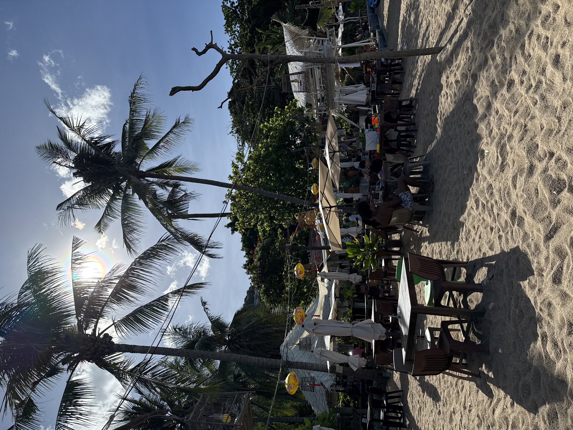 The image depicts an outdoor beachside restaurant in Baan Chaweng Noi, Thailand. Several tables and chairs are set up on the sand, some occupied by diners. Palm trees with long fronds line the background, casting shadows on the beach. In the distance, there are thatched-roof structures and lush green foliage. The sky is a clear blue with a few scattered clouds, and the sun is shining brightly, creating lens flare in the upper portion of the image. Decorative yellow lanterns hang from wires strung between poles, adding a festive touch to the scene. The overall ambiance suggests a relaxed, tropical dining experience.