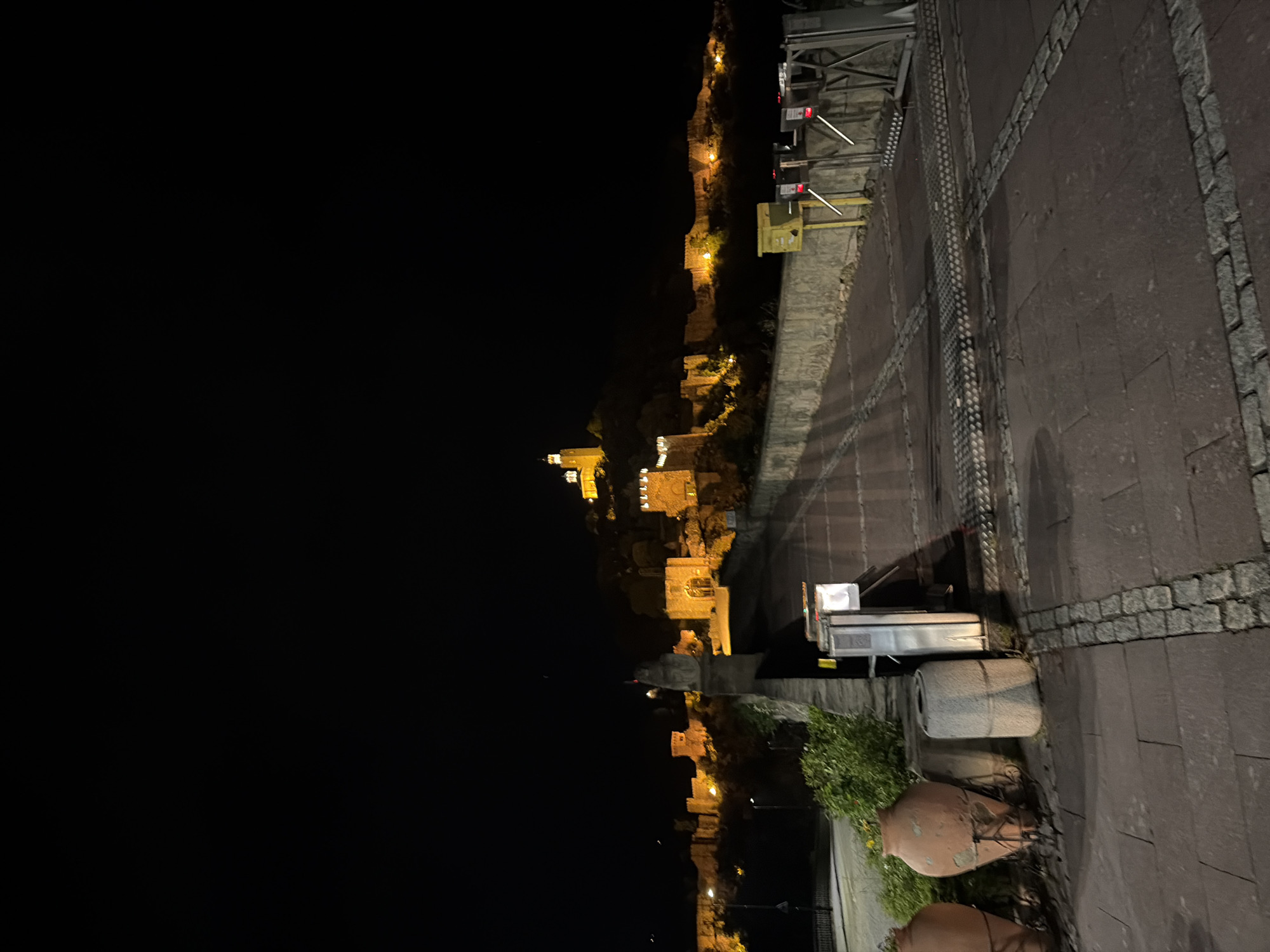 The image shows a night scene in Veliko Tarnovo, Bulgaria. A lit-up medieval fortress is visible in the distance, illuminated by numerous lights. The foreground features a stone walkway with several large, old, terracotta amphorae or similar vessels. Some green foliage grows around the base of the amphorae. A small utility cart is parked on the walkway near the vessels. Metal railings and structures are also visible, including turnstiles. The sky is dark, and the overall ambiance suggests a quiet, illuminated historic site at night.