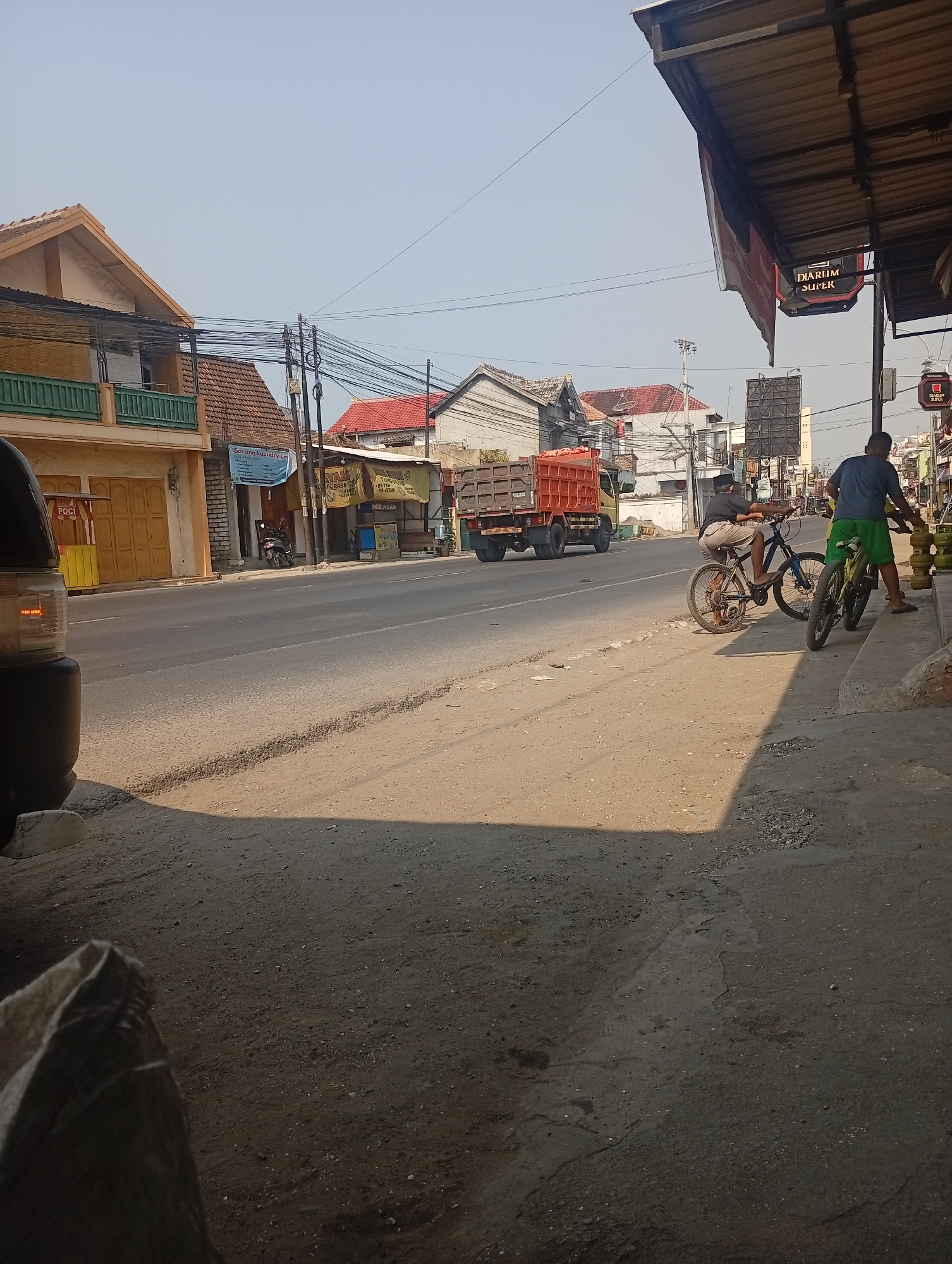 This is a street scene in Bajingjowo, Indonesia, featuring a road with a dirt shoulder. On the left side of the road, a building with a yellow facade and a green balcony is visible, adjacent to a smaller structure with yellow doors. Further down the street, several commercial buildings line the road, including one with a sign that reads "SARANG LAUNDRY EXPRESS". In the middle of the street, an orange truck is parked. On the right side of the road, two men are standing near a row of gas tanks. One man, wearing a black shirt and hat, is sitting on a bicycle. Another man, in a blue shirt and green shorts, is holding onto a bicycle. Several power lines are strung across the scene. The sky is clear and blue.