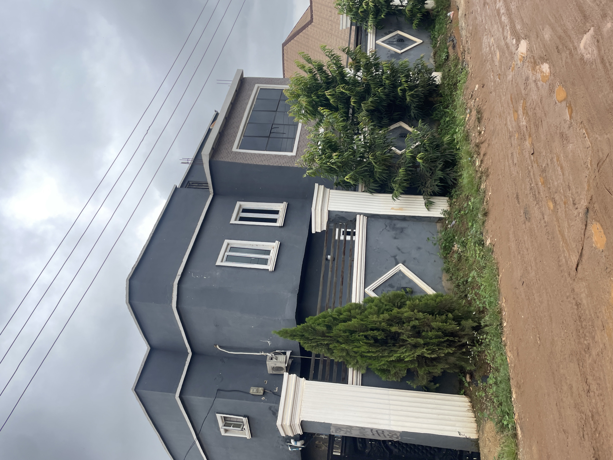 The image shows a modern, two-story house with dark gray stucco walls and white trim. The house has several rectangular windows with white frames. A large picture window with dark, reflective glass is located on the upper right side of the house. The house is surrounded by a dark gray fence with decorative diamond shapes. There are several mature green trees and bushes in front of the fence and house. A muddy, unpaved road with tire tracks and puddles is in the foreground. Power lines are visible in the overcast sky. The scene appears to be in Kurunduma, Nigeria. There are no people or animals visible in the image.