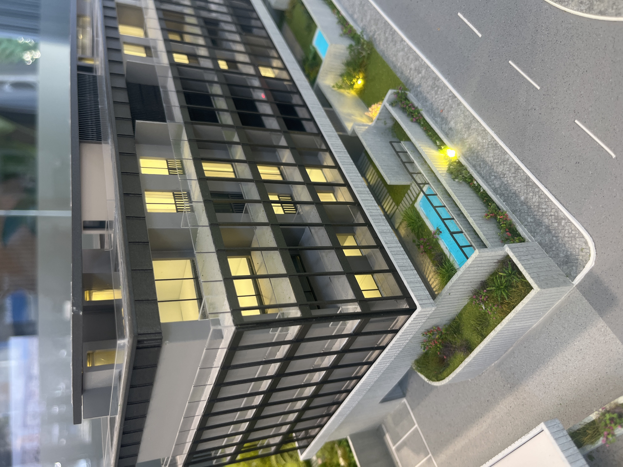 This is a close-up, eye-level, angled shot of an architectural model of a modern, multi-story building. The building features a dark grey facade with large, black-framed windows that illuminate with a warm yellow light, suggesting interior lighting. The upper levels have balconies with clear glass railings and some sections of grey paneling. The top of the building is flat with some darker grey ventilation structures.
At the base of the building, there is a landscaped area with artificial grass, small flowering plants in shades of pink and red, and two small, bright blue rectangular swimming pools. Concrete retaining walls and pathways surround the landscaped areas and pools. A grey asphalt road with white dashed lines runs along the right side of the model. The overall impression is a model of a residential or commercial building, likely a condominium or hotel, designed with contemporary aesthetics. There are no people or animals visible in the scene. The backdrop is blurred, suggesting it is either a display environment or a reflection of other objects. The lighting is bright and even, highlighting the details of the model.