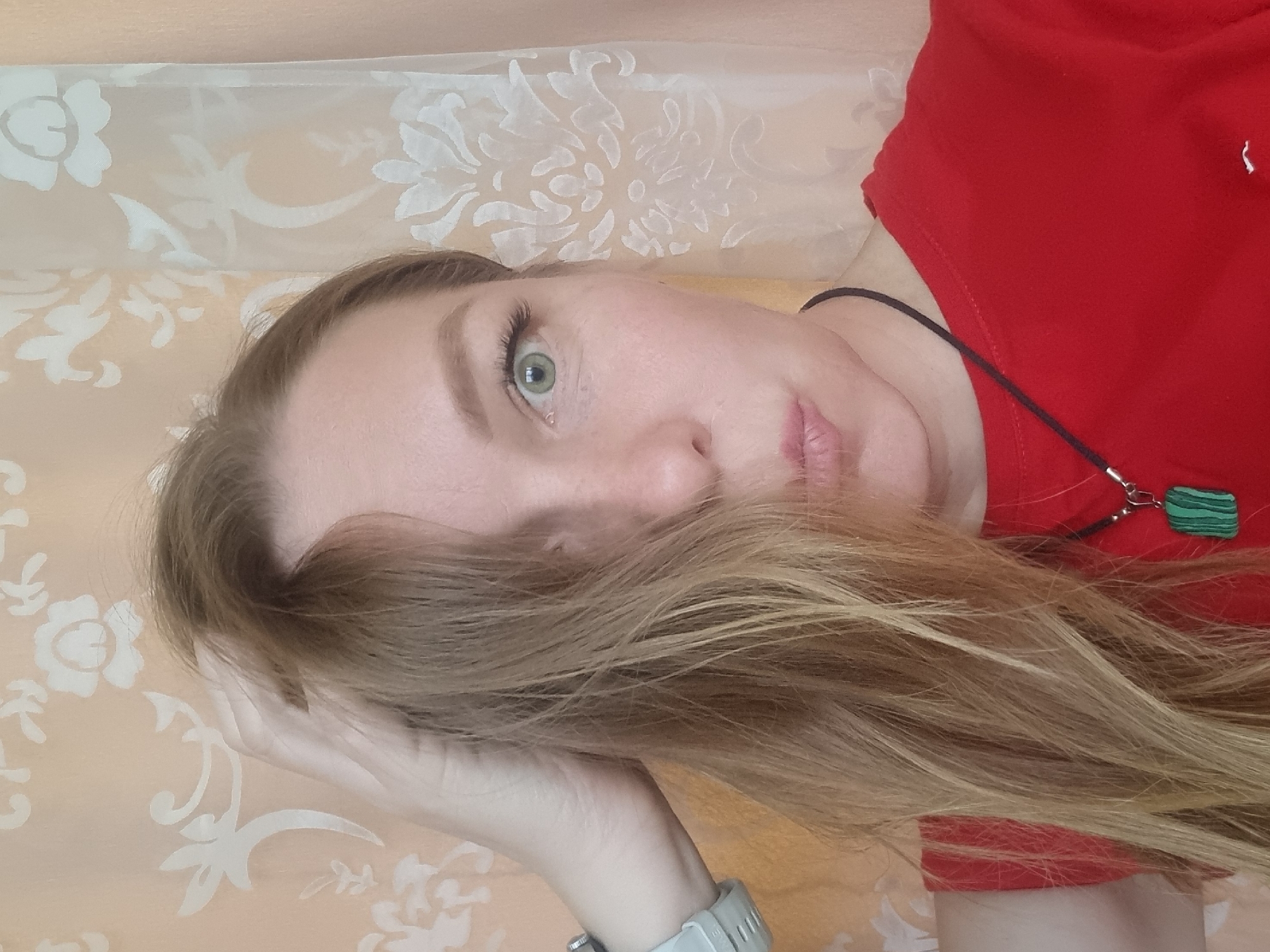 A portrait of a fair-skinned woman with long, blonde hair. The woman is reclining, with her head resting on her hand. She is wearing a red shirt and a necklace with a green pendant. Her eyes are green. The background is a floral-patterned peach-colored material, possibly a curtain or wall covering. The image does not provide any information to infer a location in the city of Chaikovsky, Russia.