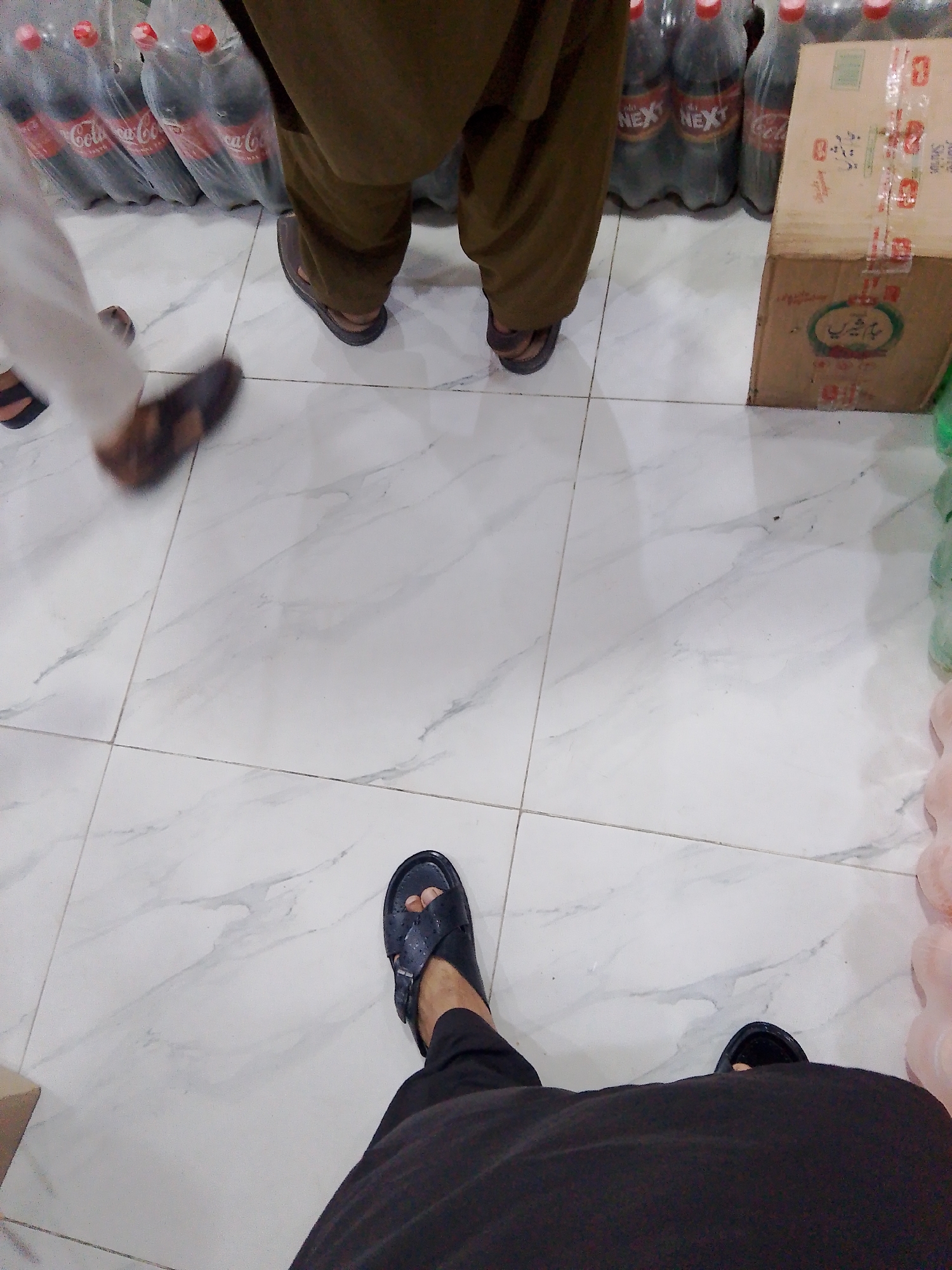 The image depicts a ground-level view of a retail setting, likely a small shop or market stall, in Mansehra, Pakistan. The floor is tiled with a white marble pattern. In the background, multiple large plastic bottles of Coca-Cola and what appears to be "Next" brand beverages are stacked. To the right, a cardboard box with packaging tape and some lettering is visible, possibly indicating merchandise. A person's legs are visible in the foreground, wearing black trousers and black sandals. Another person's legs and feet are also visible on the left, with one foot wearing a brown sandal and the other in motion, wearing a dark sandal. A white garment is also visible on the person on the left. The scene suggests a commercial environment where beverages are sold.