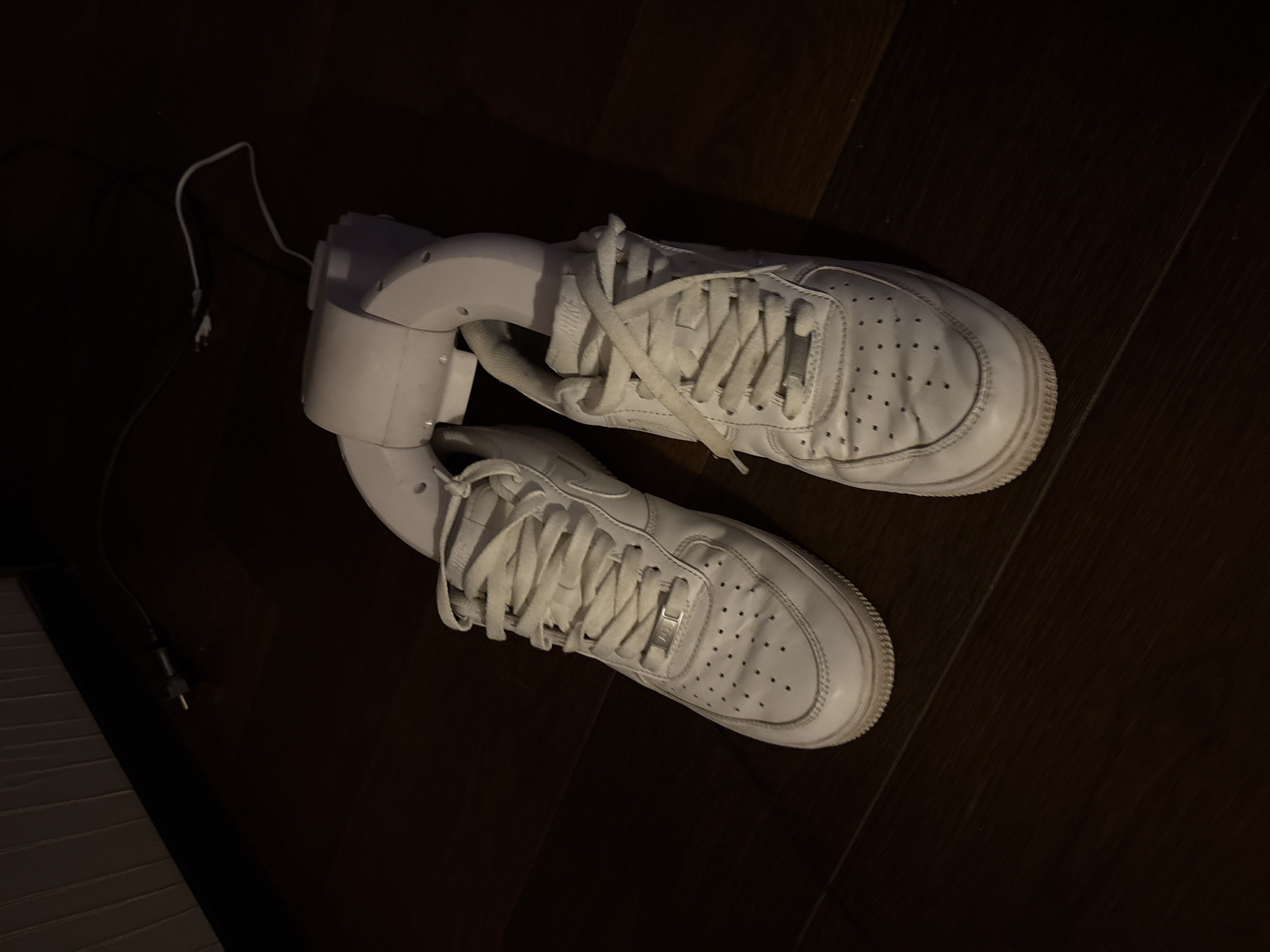 The image shows a pair of white Nike Air Force 1 sneakers, with white laces. The sneakers are connected by a white, curved electronic device, possibly a shoe dryer or deodorizer, which has a visible power cord. The scene is set on a dark wooden floor. There is no indication of the city of Ореховка, Russia in the image.