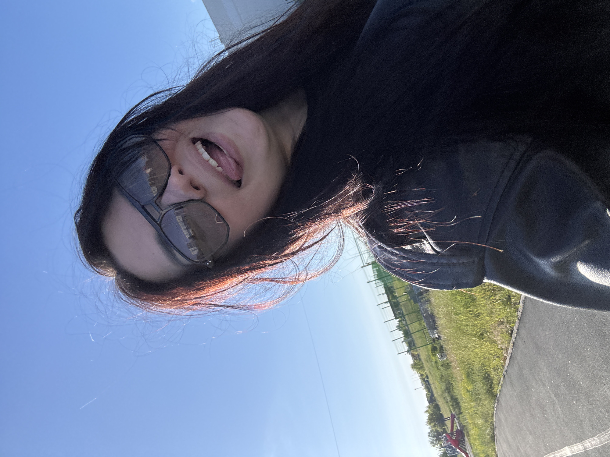 An outdoor, brightly lit image, strongly tilted to the right, features a person in a playful pose. The subject, visible from the chest up, has long, dark hair, with some strands appearing reddish-brown where sunlight catches them. They are wearing dark sunglasses and a black jacket or top. Their mouth is open, revealing teeth and a slightly extended tongue, suggesting a lighthearted or expressive mood.
The background is dominated by a clear, vibrant blue sky that occupies most of the upper left portion of the image. The lower right reveals a paved path, likely asphalt, bordered by a strip of green grass. Beyond the grass, a fence-like structure and what appear to be distant trees or other landscape features are visible. The bright sunlight indicates it is daytime, likely midday or early afternoon, with clear weather. The setting appears to be a park, a walking trail, or an open recreational area. No visible text is present in the image.