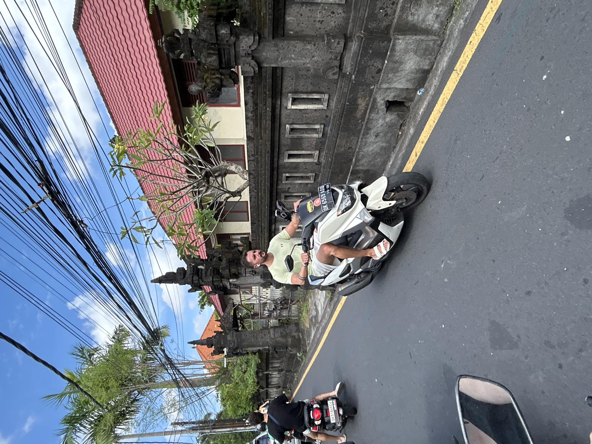 A man is riding a white scooter down a street in North Kuta, Indonesia. He is wearing a light green t-shirt and white shorts, with white sandals. He has a beard and is looking forward. Behind him, another person on a black scooter is visible. The street is paved with asphalt and has a yellow line painted along the side. A stone wall with decorative carvings lines the street, and beyond that are buildings with red tile roofs and palm trees. Overhead, a dense network of power lines is strung across the sky. The sky is blue with scattered clouds.
