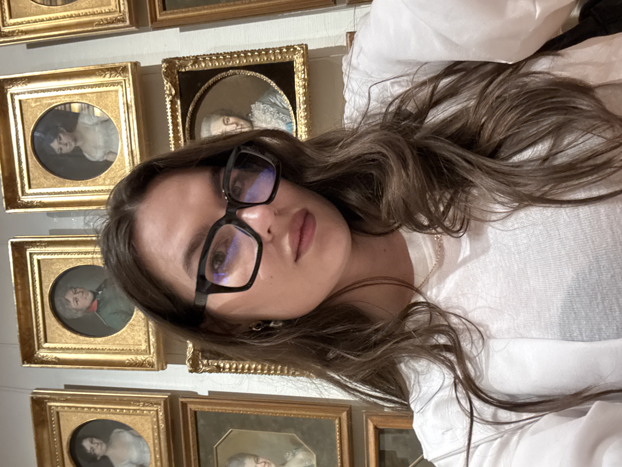 A woman with brown hair and glasses is posing for a selfie in front of a wall covered in framed portraits. She is wearing a white shirt and a gold chain necklace. The portraits appear to be miniatures in ornate gold frames. The location is likely a museum or gallery in Moscow, Russia, considering the type of artwork displayed.