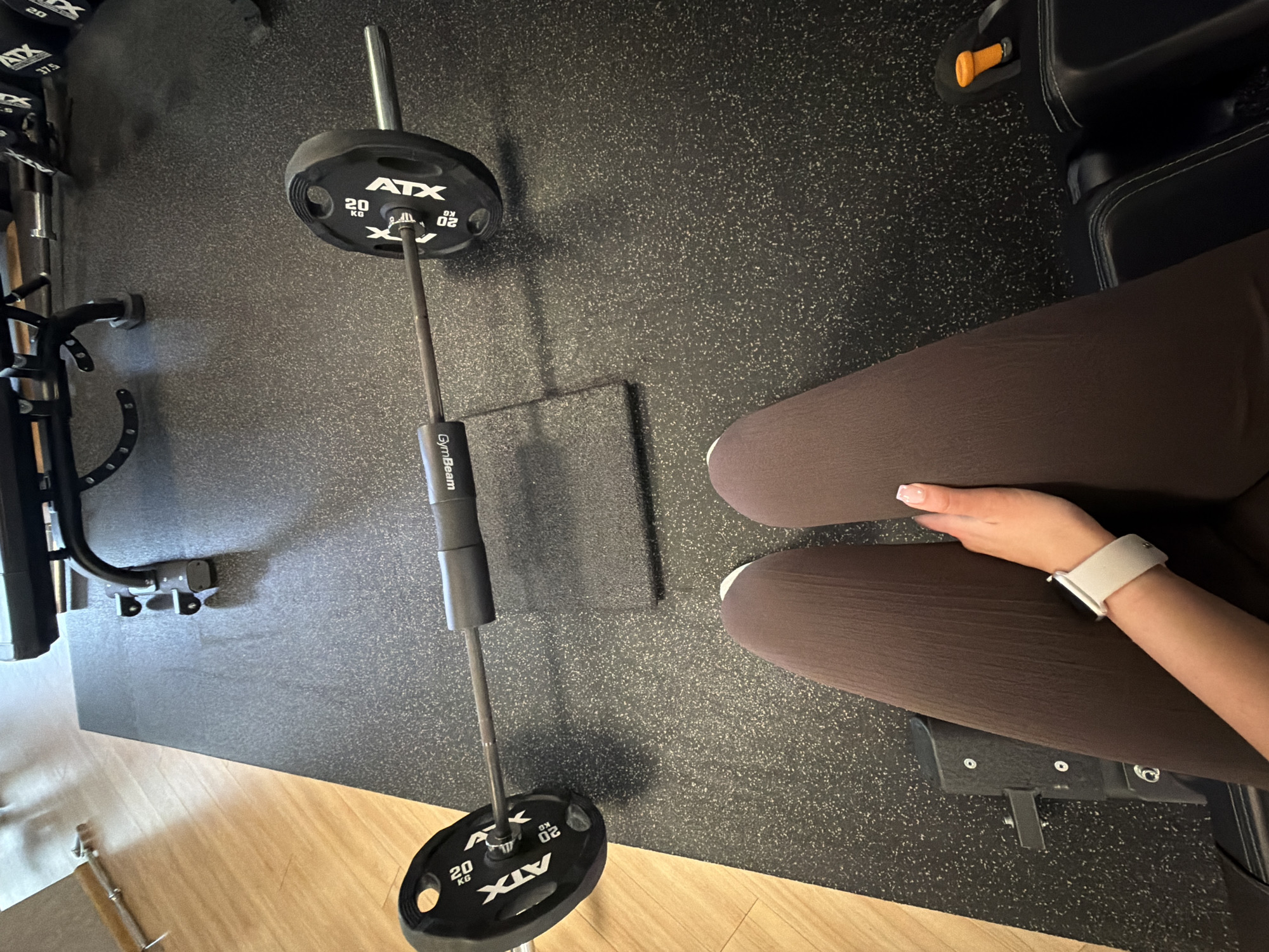 The image shows a first-person perspective of someone sitting at a gym. In the foreground, brown leggings and a hand with a smartwatch are visible. The gym floor is covered with black rubber mats. A barbell with 20kg ATX weight plates on each side is positioned horizontally in the middle of the frame. In the background, a weight bench and other gym equipment are partially visible. There is no information to suggest the location is in Gabrovo, Bulgaria.