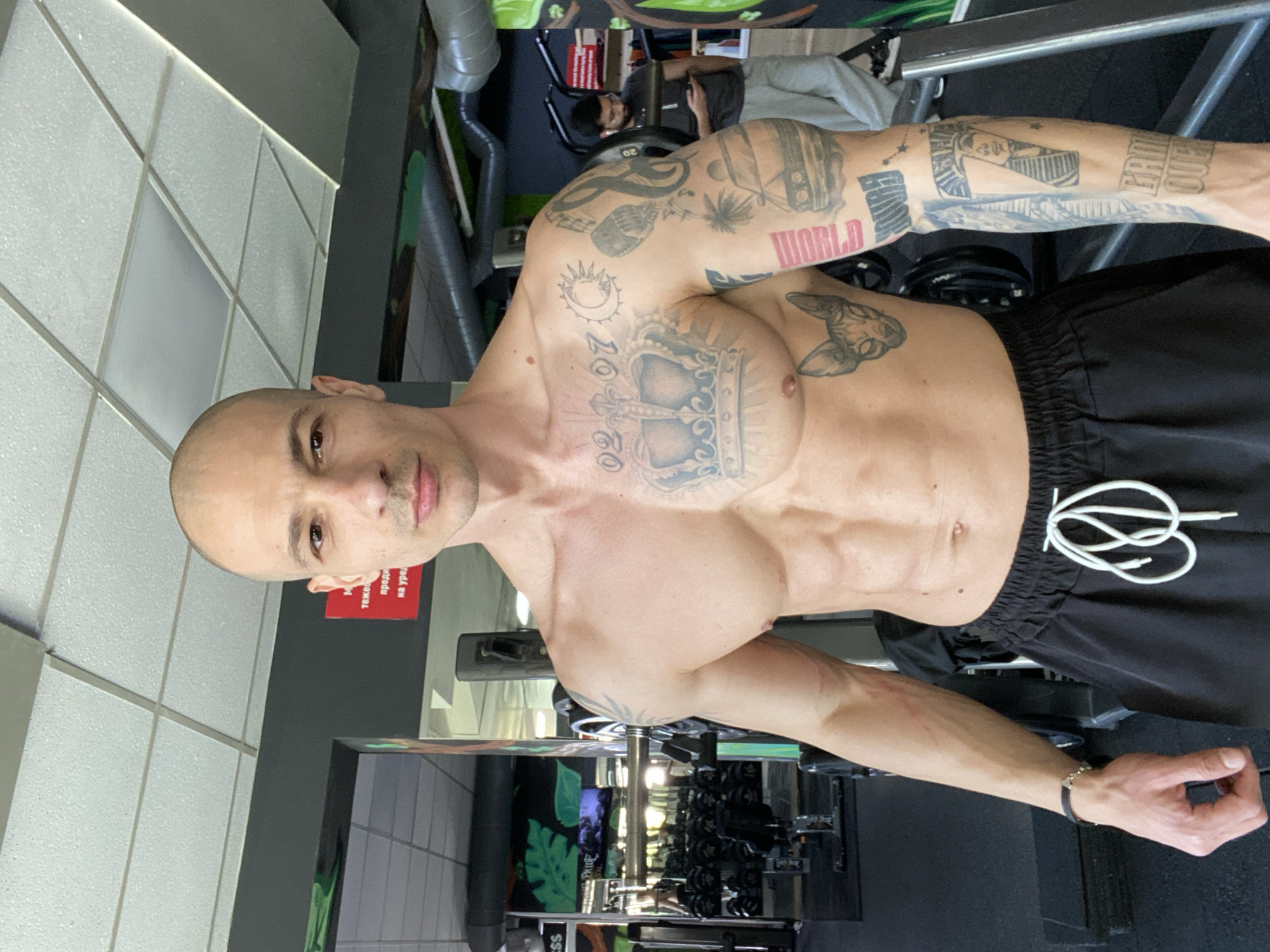 A shirtless muscular bald man with tattoos is posing in a gym. He is wearing black shorts with white drawstrings. The gym has various weight training equipment and dumbbells. The gym is located in the city of Pleven, Bulgaria. Another person is visible in the background, using a machine.