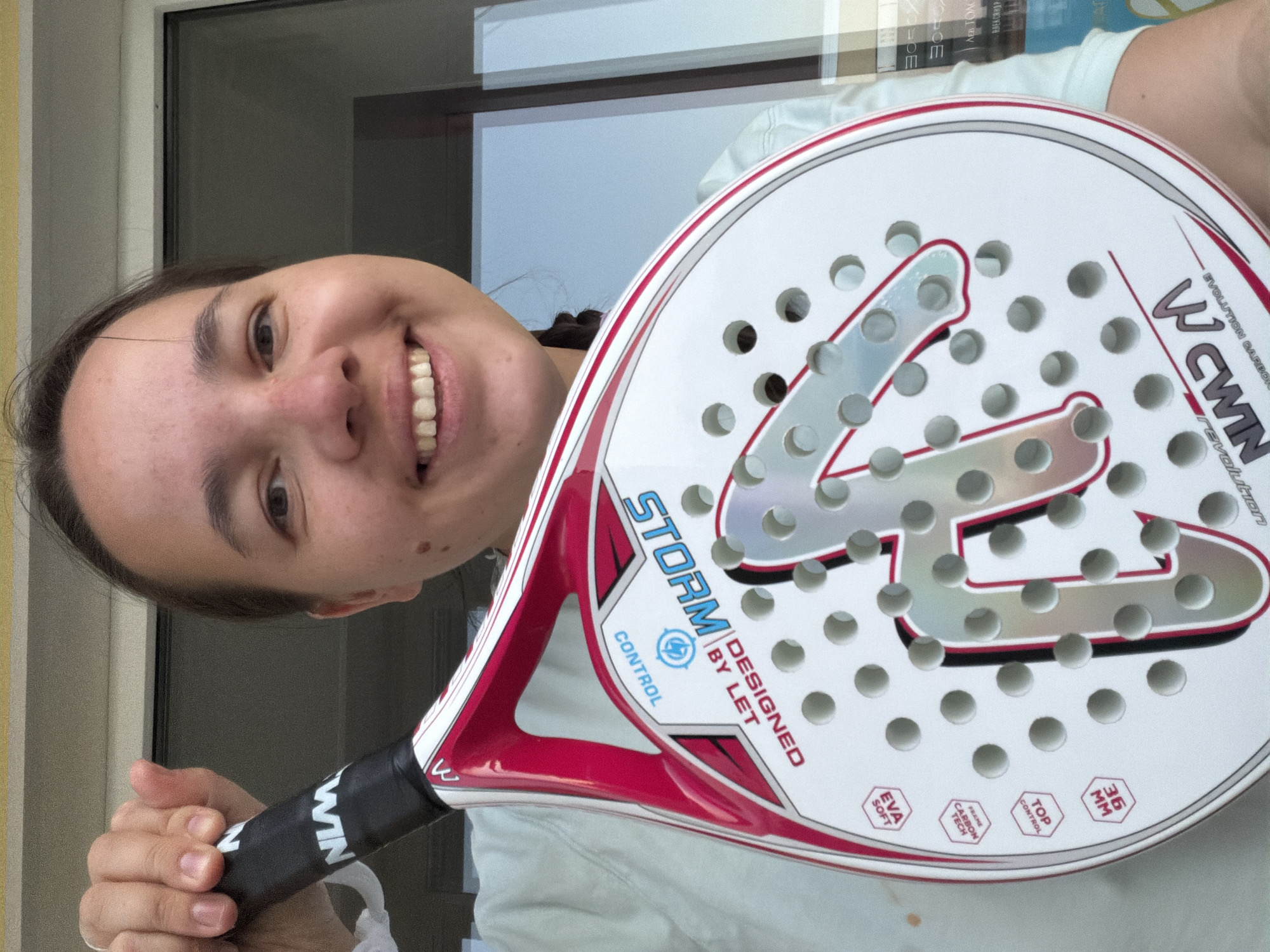 A young woman is holding a white and red WCWIN Storm padel racket, angled to display its features. The racket has numerous holes in the face and text indicating "EVOLUTION CARBON revolution," "STORM," "DESIGNED BY LET," "CONTROL," "EVA SOFT," "FRAME CARBON TECH," "TOP CONTROL," and "36 MM." The woman is smiling and looking at the camera. Behind her is a window and some bookshelves. There is no evidence suggesting this picture was taken in Chaikovsky, Russia.