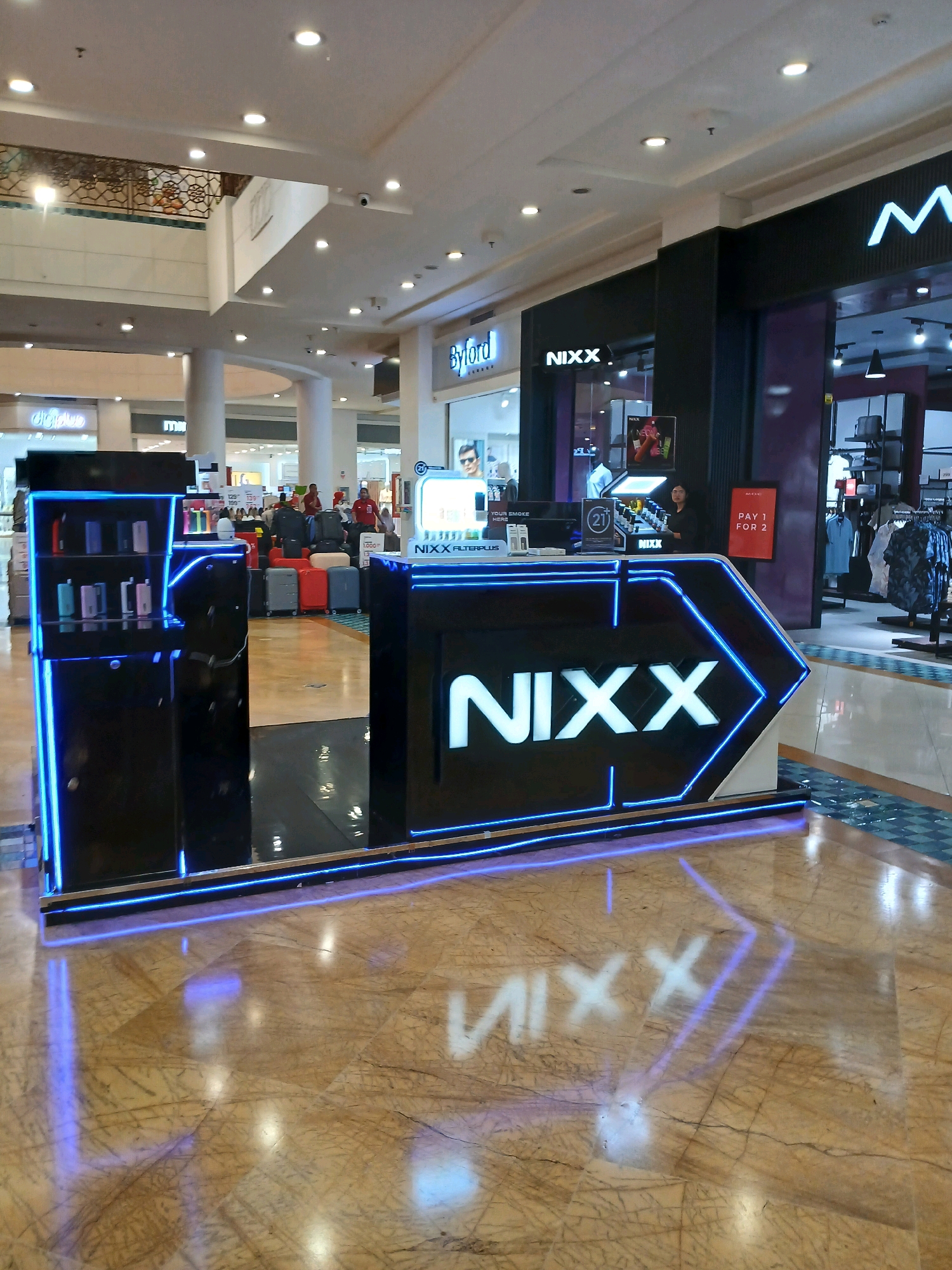 The image shows a NIXX branded kiosk in a shopping mall. The kiosk is illuminated with blue neon lights and displays various products on shelves. The kiosk has a large "NIXX" logo on its front. In the background, there are other shops and shoppers. The mall appears to be modern with marble flooring and a high ceiling. The location of this mall is in North Jakarta, Indonesia.