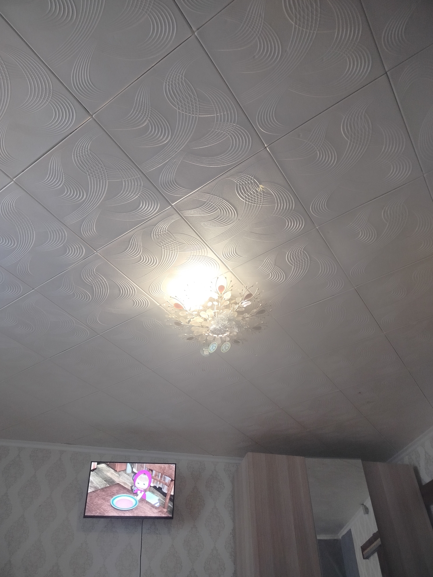 The image captures an indoor scene, likely a room in a residential building, with a view of the ceiling and a portion of the wall. The ceiling is made of white tiles with a subtle, repetitive wavy pattern. A modern chandelier with a floral-like design and multiple light sources hangs from the center of the ceiling, illuminating the area directly below it. To the left of the frame, a flat-screen television is mounted on the wall, displaying a cartoon image of a character named Masha from the animated series "Masha and the Bear." The television is positioned above a patterned wallpaper. On the right side of the image, a section of a wooden wardrobe with a mirrored door is visible, reflecting a part of the room. The overall environment appears to be domestic. The image was taken in the city of Часцы, Russia.