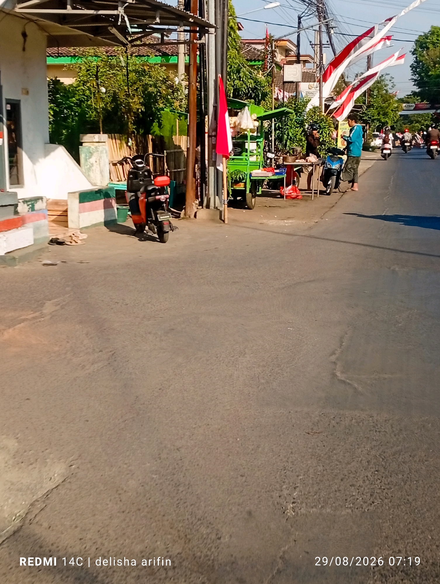 In Plinggisan, Indonesia, a street scene unfolds with a bright, sunny atmosphere. The image captures a section of a road with a building on the left and trees lining the right side. Several people are visible. Two individuals are standing near a green food cart. One person, wearing a blue jacket and dark pants, is interacting with another person who appears to be standing behind the cart. A blue scooter is parked in front of them. Further down the street, more people are seen on scooters, suggesting ongoing activity. Red and white Indonesian flags are prominently displayed, some fluttering in the breeze. An orange scooter is parked to the left of the food cart. The overall scene depicts a typical day on a street in a populated area, with everyday activities and local decorations.