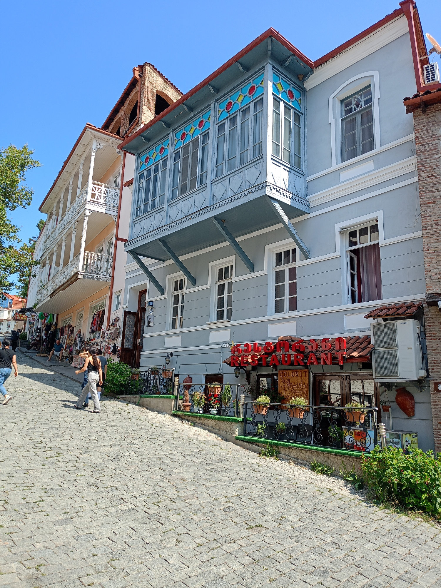The image depicts a street scene in Sighnaghi, Georgia, on a clear, sunny day. The foreground shows a cobblestone street with several people walking. Two individuals, appearing to be women, are the most prominent. One, wearing a black top and blue jeans, walks away from the camera to the left. Another, in a black tank top and light-colored pants, walks towards the right, her back to the camera.
The background is dominated by the architecture of Sighnaghi. On the right, a light gray building features a blue, ornate balcony with stained glass windows above a restaurant with a red neon sign that reads "RESTAURANT." An air conditioning unit is mounted on the exterior wall to the right of the restaurant entrance. Next to this building, a section of a brick wall is visible.
To the left of the gray building, a narrower building with a white ornate balcony extends upwards. This building appears to be a hotel or guesthouse, with several floors and balconies. Shops are set up along the sidewalk in front of this building, displaying various goods. A green tree partially shades the scene on the far left. The overall impression is a picturesque street in a historic Georgian town.