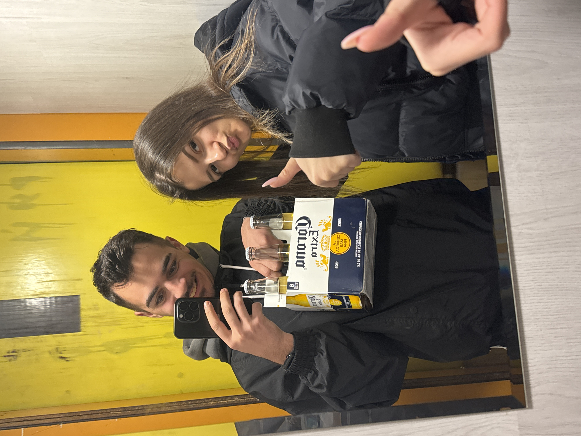 A mirrored selfie captures a man holding a six-pack of Corona beer in front of a yellow wall with orange trim. He holds a black smartphone, presumably taking the picture. To the right of the frame stands a woman with long brown hair wearing a black puffy jacket. She points at the beer. The mirror's reflection shows a light-colored floor and what appears to be a border surrounding the mirror itself. It's not possible to determine the specific location in Sofia, Bulgaria based on the image alone.