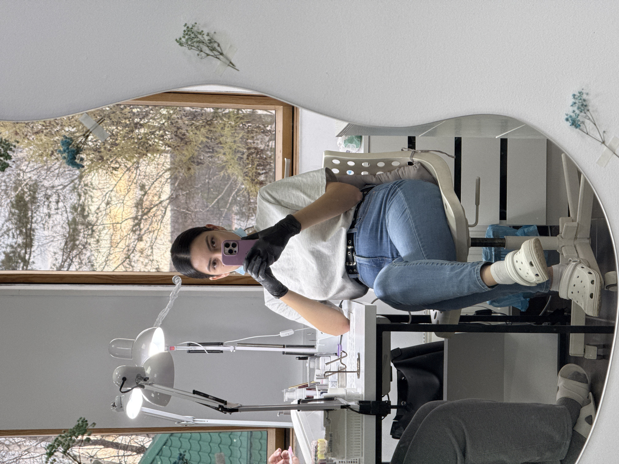 A young woman is captured in a mirror selfie, posing in what appears to be a beauty or nail salon setting. The image is taken at an angle, with the woman looking into her purple smartphone as she holds it up to capture her reflection. She has dark hair pulled back and is wearing a light-colored t-shirt, blue jeans with a black belt, black gloves, and white Crocs.
The mirror she's using has a distinctive wavy top edge and is partially obscured by her body. In the reflection, behind her, are two bright, articulated desk lamps illuminating a white table covered with various small containers, brushes, and tools, suggesting nail or lash artistry equipment.
To the woman's left (in the reflection), a large window offers a view of a bare deciduous tree outside, implying a cold season like late autumn or winter. Bits of dried foliage, both green and blue, are decoratively taped to the white wall above and to the side of the mirror.
Another person's legs, clad in grey pants and white slides or slippers, are visible in the lower part of the mirror's reflection, suggesting a client seated opposite the woman. The overall scene conveys a professional yet casual workspace. There is no discernible text visible in the image.