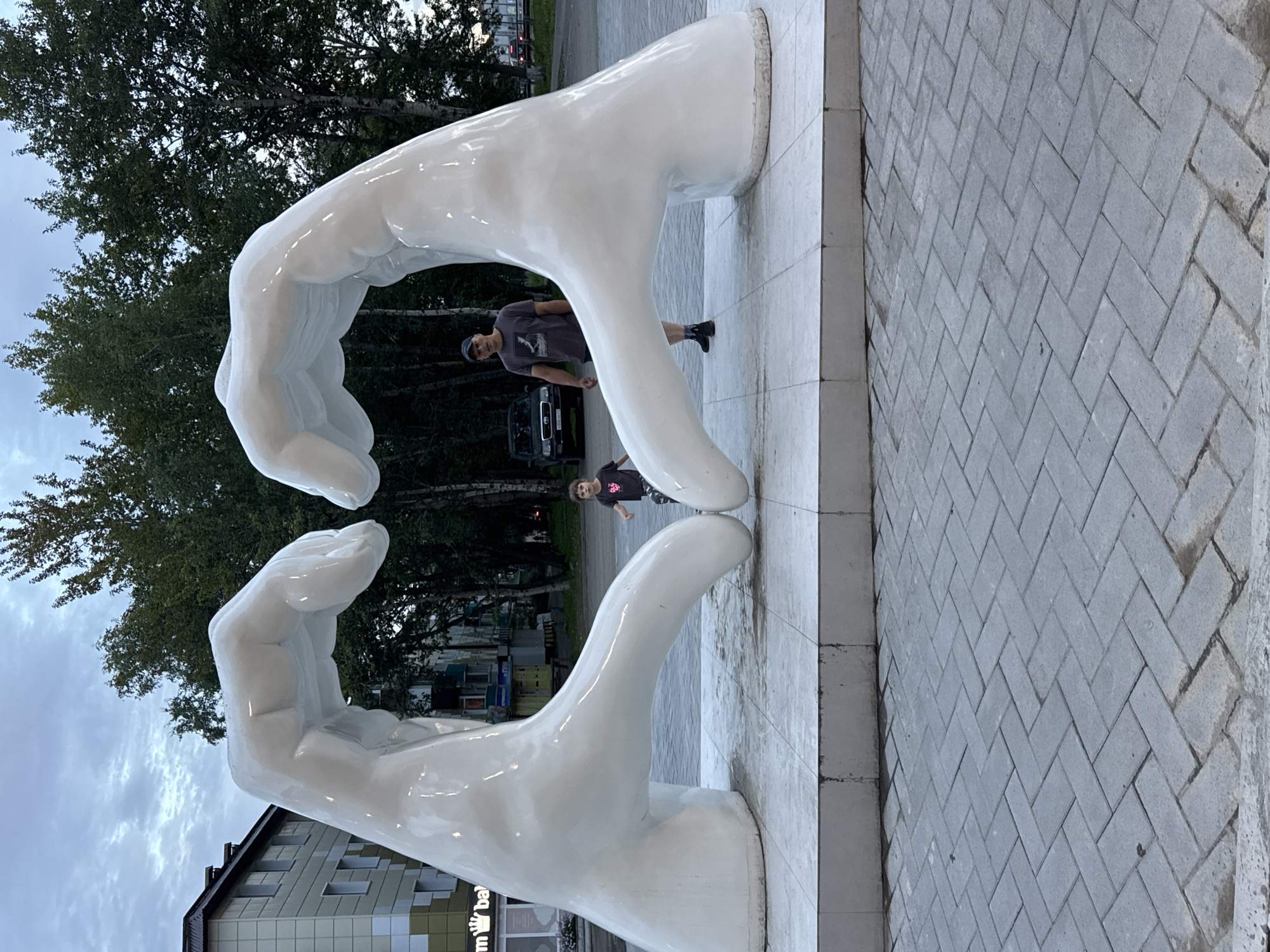 This image depicts a large, white sculpture of two hands forming a heart shape. A man wearing a dark t-shirt and a baseball cap stands to the right of the sculpture, looking towards the camera. A young child is positioned between the sculpture's hands, facing the camera and appearing to be running. The sculpture is situated on a paved surface in front of buildings and trees, suggesting an outdoor urban environment. The sky is overcast. This sculpture is located in the city of Yelizovo, Russia, as indicated by the "m ba" logo on a building in the background, which is a partial view of the "m-ba" shopping center, a landmark in Yelizovo.