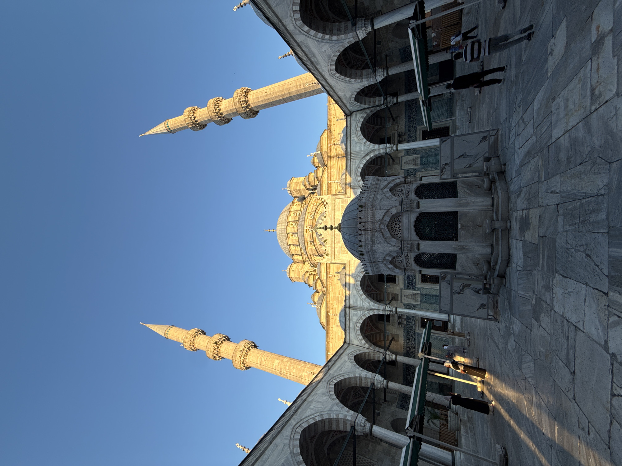 This is a low-angle, wide shot of the courtyard of the New Mosque (Yeni Camii) in Istanbul, Turkey, taken during the late afternoon or early evening, as indicated by the long shadows cast by the setting sun. The mosque's grand facade, featuring a central dome and multiple smaller domes and minarets, dominates the background under a clear, deep blue sky. Two prominent, slender minarets flank the main structure, their intricate stone carvings illuminated by the warm light. In the foreground, a marble courtyard is paved with large, irregular flagstones. An ornate, multi-tiered fountain structure sits in the center of the courtyard. Along the sides of the courtyard, a covered walkway with arched colonnades, made of white marble, extends from the main building. Several individuals are present in the courtyard. On the left, two people in traditional Islamic attire, one in black and the other in white and black, are walking away from the camera, their figures elongated by the strong shadows. Further back, a few other figures can be seen strolling through the courtyard or standing. Green awnings are visible along parts of the walkway, providing shade. The overall scene conveys a sense of architectural grandeur and peaceful contemplation.
