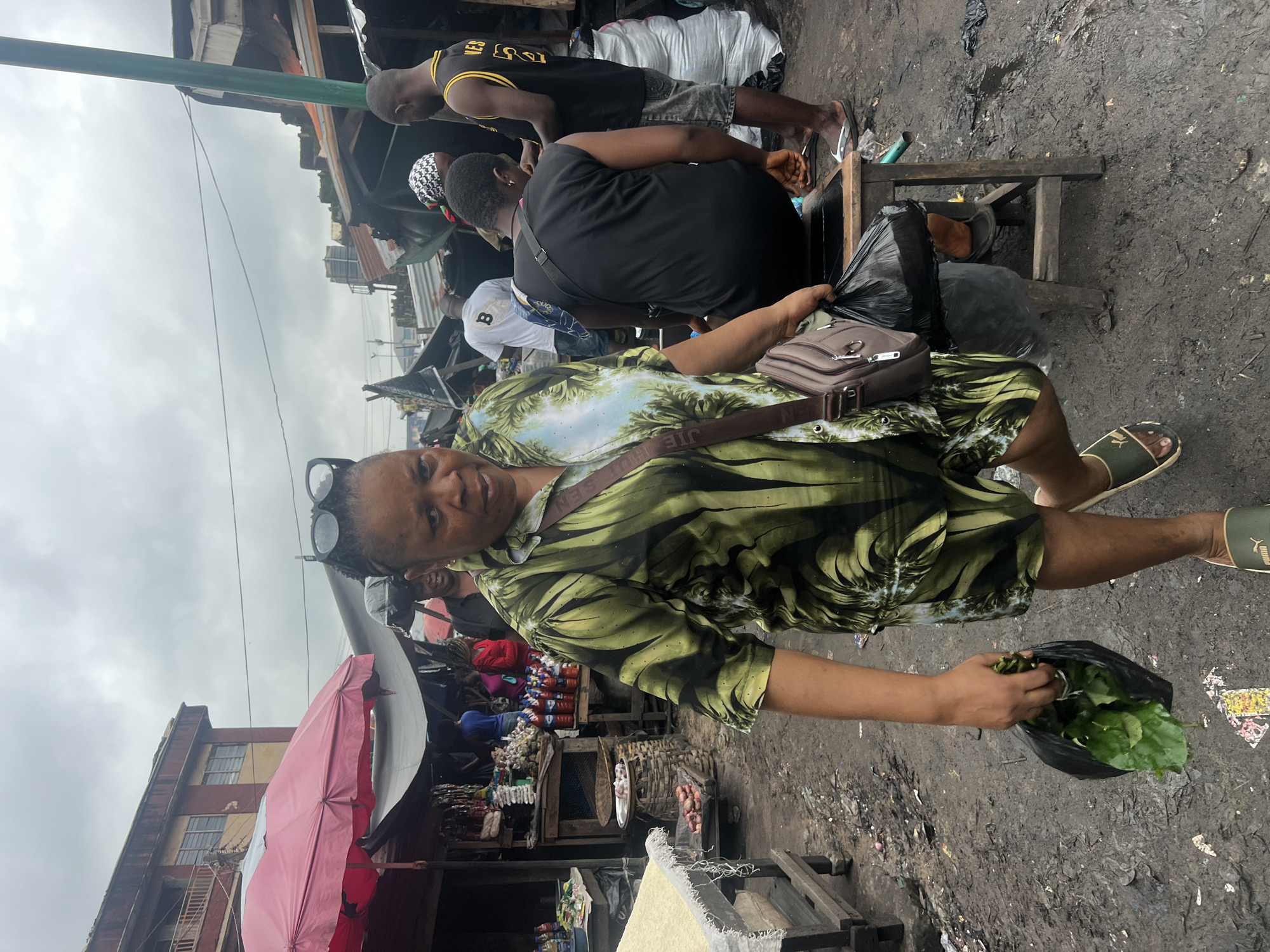 The image depicts a busy outdoor market scene in Nkpor, Nigeria. A woman in the foreground, wearing a green patterned shirt and shorts, walks towards the camera, carrying a black plastic bag filled with leafy green produce in her left hand and another black plastic bag in her right. She has sunglasses perched on her head and a brown crossbody bag over her left shoulder. The ground is muddy and littered with debris, indicating a typically unpaved and well-trodden market environment.
Behind her, several other people are visible, some engaged in transactions or waiting. A man in a black tank top with yellow lettering stands to the right, facing away from the camera. To his left, a person in a black sleeveless top sits on a wooden stool, also with their back to the camera. Several market stalls with corrugated metal roofs and awnings are set up, displaying various goods including bottles of drinks and what appear to be produce and packaged items. The overall atmosphere suggests a typical day at a bustling Nigerian market. The sky is overcast.
