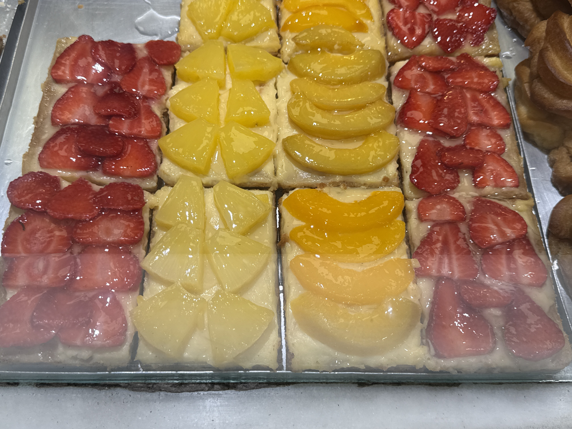 The image displays a clear glass-fronted bakery display case containing multiple rectangular fruit tarts on a metallic tray, located in the City of Unknown, Unknown. The tarts are arranged in four horizontal rows and two vertical columns. Each tart features a pastry base topped with a light-colored cream or custard, uniformly glazed with various fruit toppings. The top row consists of tarts topped with sliced strawberries. The second row displays tarts with triangular-cut pineapple pieces. The third row contains tarts adorned with sliced peaches. The bottom row mirrors the top row with sliced strawberry toppings. All fruit toppings appear shiny due to a glaze. Below the bottom row of tarts, blurred pastry items, possibly croissants, are visible. The left side of the frame shows the thick edge of the transparent display glass and a white surface, indicating an indoor display setting. No individuals are present in the image.