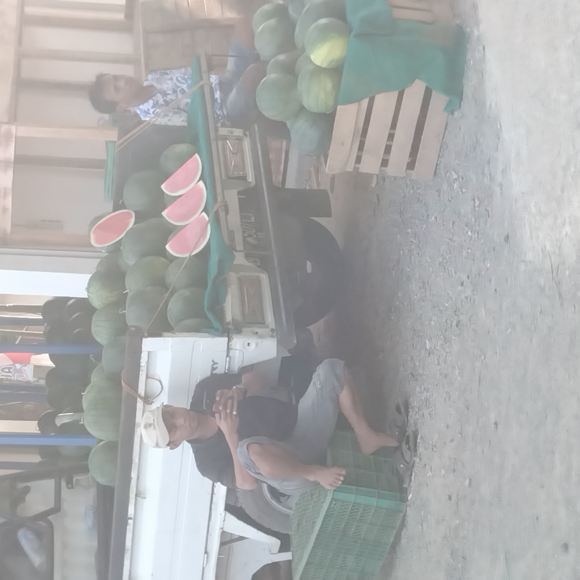 The image captures an outdoor scene, likely a market or roadside stall, in Pelaihari, Indonesia, indicated by the license plate "501 LJ". In the foreground, a man wearing a black t-shirt and grey shorts sits on a green plastic crate, looking at his smartphone. He is barefoot and wearing sandals on his left foot. Behind him, a white utility truck is loaded with numerous watermelons. The truck bed displays several whole watermelons, and to the right, slices of watermelon are arranged on a green cloth. Further to the right, another man, wearing a white shirt with a blue floral pattern, stands near the watermelons. The ground is covered with gravel and dirt. In the background, wooden structures and a glimpse of a flag suggest a commercial or communal area. The overall lighting is diffused, possibly due to hazy conditions.