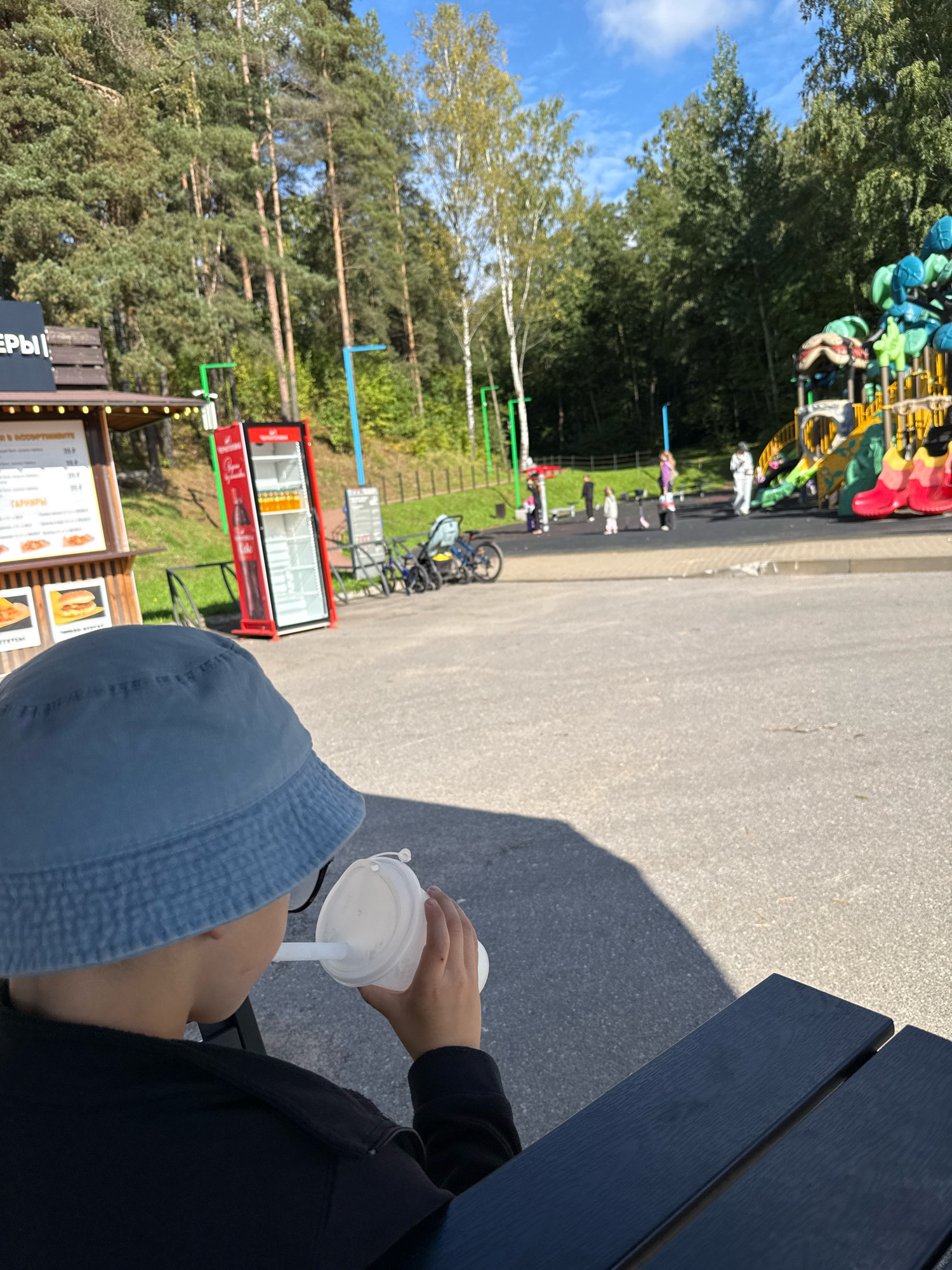 The image depicts an outdoor scene at what appears to be a recreational area or park, likely in Vsevolozhsk, Russia, given the provided context. In the foreground, a child wearing a blue bucket hat and a black jacket is seated at a dark blue picnic table, drinking from a white disposable cup with a straw. The child's face is partially obscured, but their arm and hand are visible holding the cup. The background features a playground with slides and climbing structures, some of which are brightly colored. Several children and adults are scattered around the playground area. To the left of the playground is a food stand with menus and a red refrigerator. The ground is paved with asphalt and there are trees and foliage surrounding the area, indicating a natural setting. The sky is blue with scattered clouds. The overall scene suggests a sunny day at a family-friendly location.