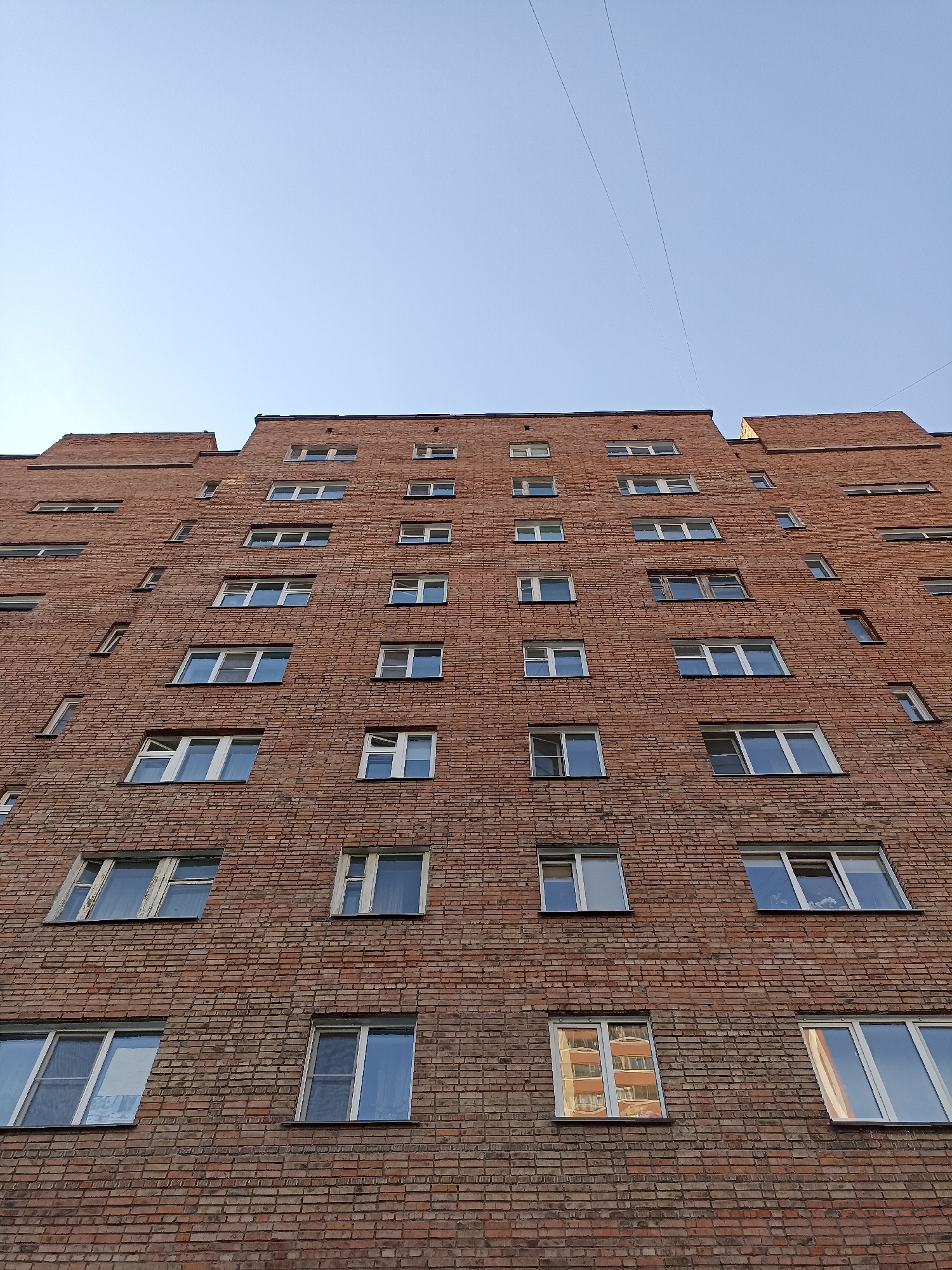 This is a low-angle, full shot of a brick apartment building in Novosibirsk, Russia, under a clear blue sky. The building is tall and appears to be constructed from red bricks. It has numerous windows, most of which have white frames and appear to be closed. Some of the windows reflect the sky, while others show faint interior lights or patterns of adjacent buildings. There are two thin power lines stretching across the upper portion of the frame, against the sky. The architecture suggests a Soviet-era or post-Soviet residential building. There are no people visible in the frame.