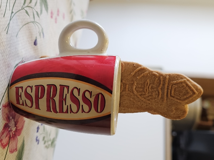 A vertical close-up photograph captures a Sinterklaas-shaped gingerbread cookie positioned upright within a red and brown ceramic mug. The mug, bearing the word "ESPRESSO" in cream-colored capital letters within an oval design, has a white handle visible on its left side. The light brown cookie displays an embossed relief depicting a figure, likely a bishop or Saint Nicholas, complete with a miter and facial details. The mug and cookie rest on a white tablecloth patterned with stylized red/pink flowers and green leaves. The background is blurred, showing light-colored walls or surfaces and an indistinct dark object, possibly part of an appliance or furniture. The scene is set indoors, likely in a kitchen or dining area, in Esch-sur-Alzette, Luxembourg.