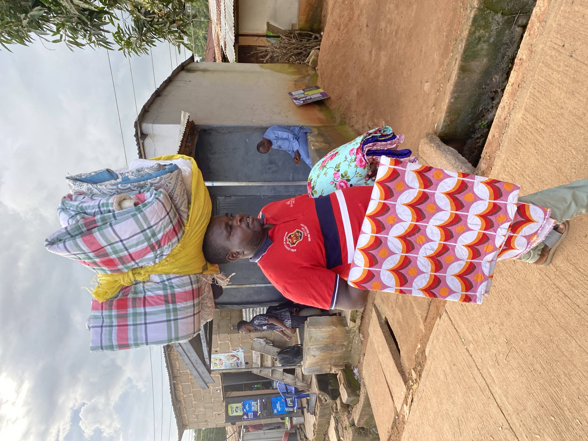 A man in Bukuru, Nigeria, balances a large load of folded textiles on his head. He is wearing a red polo shirt with navy and white trim and green trousers. In his right arm, he carries more folded fabric, one piece patterned with pink, orange, and white circles, another with a floral print. The man stands on a concrete path in front of a row of buildings that appear to be shops or residences. The buildings are constructed from concrete blocks and corrugated metal roofing. In the background, a man in a blue shirt sits by an entrance, and another younger man stands near a staircase. The sky is overcast with scattered clouds.