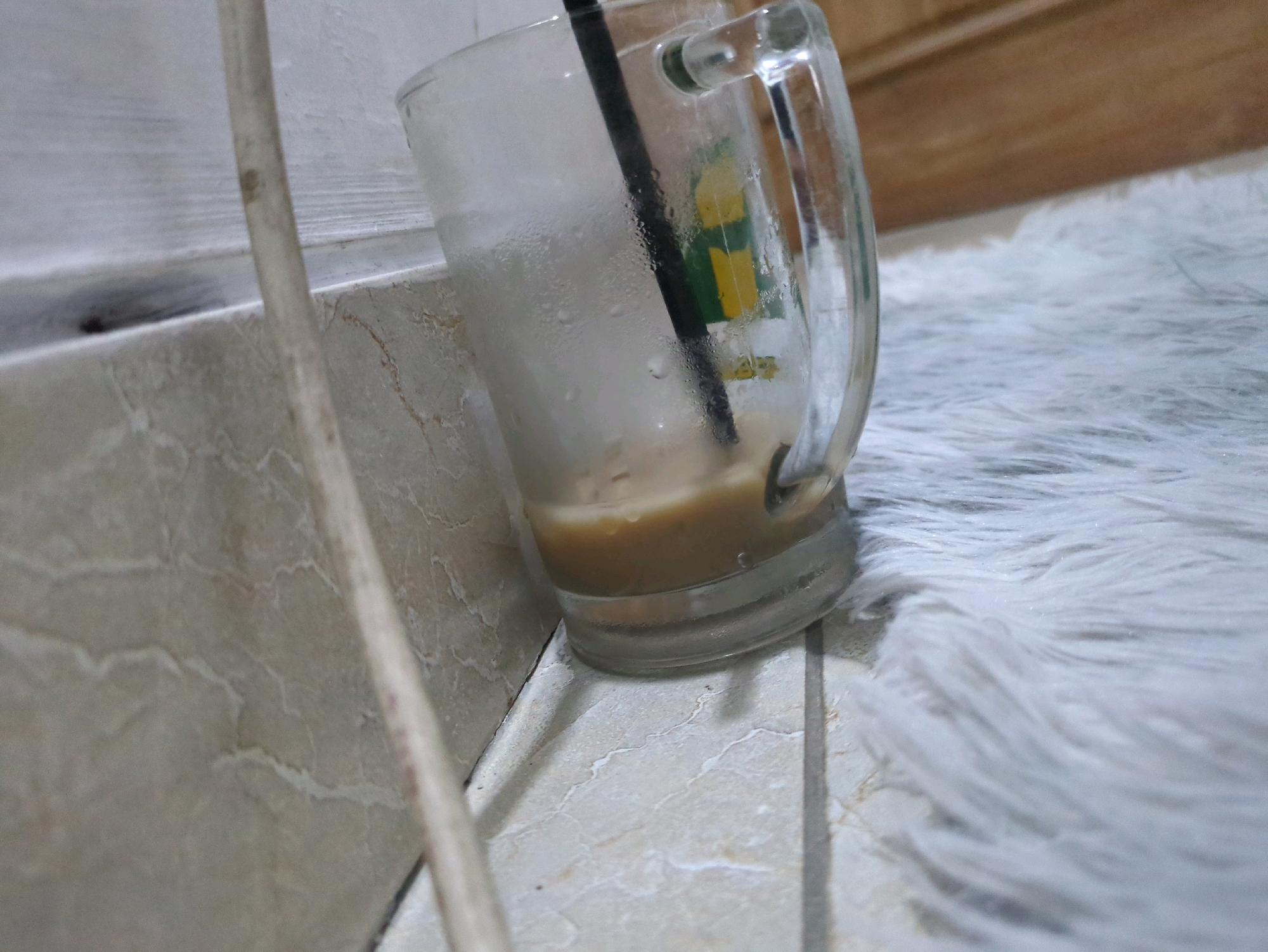 An iced coffee drink is in a glass mug that is covered in condensation. The mug has a handle and a black straw. The mug sits on a tiled surface, and next to it is a fluffy white rug. In the background, there is a wooden surface and a white wall. The scene is indoors, likely in a home in Sindon, Indonesia.