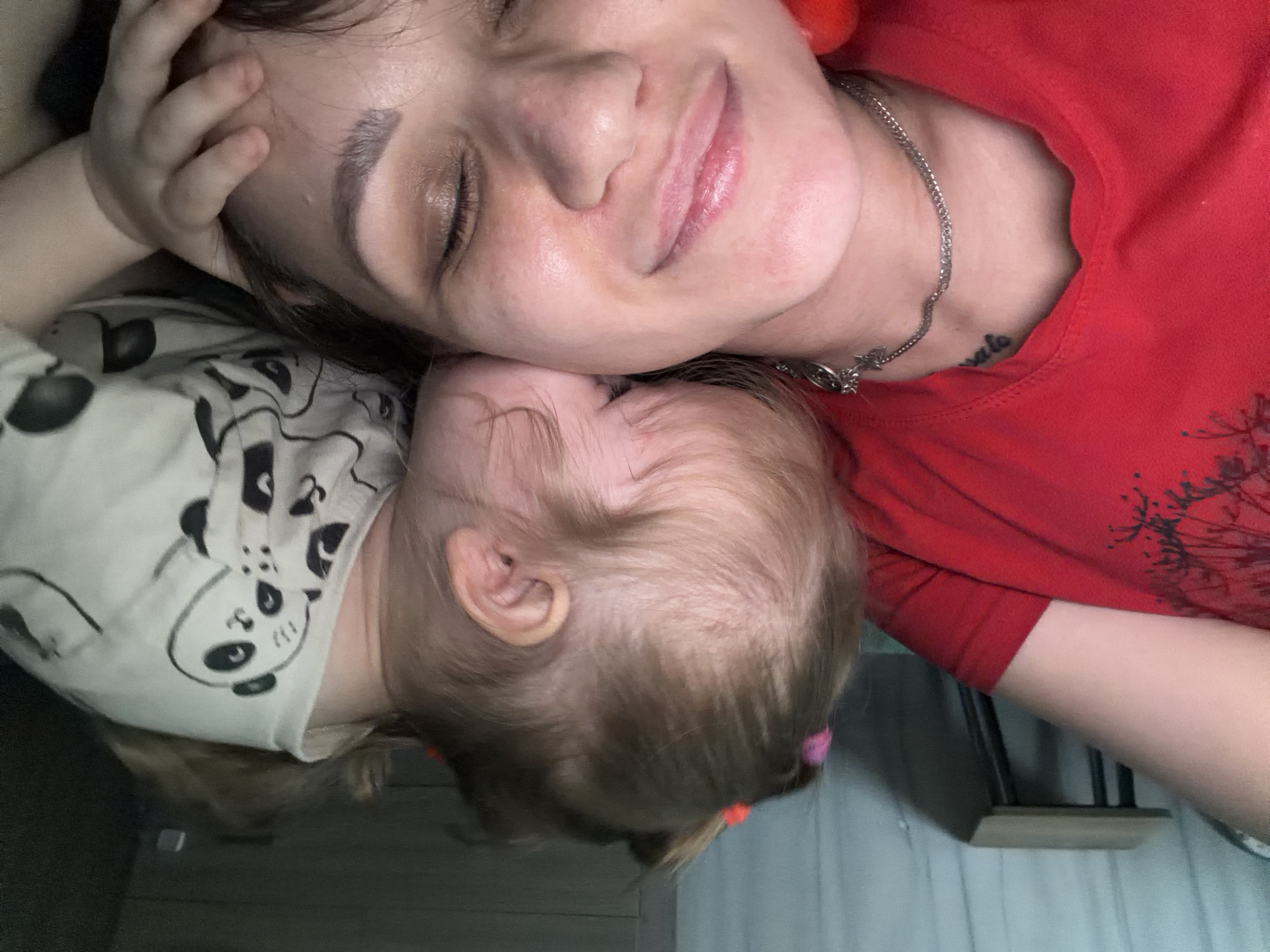 The image depicts an intimate, overhead view of an adult female and a young child in an indoor domestic setting. The adult female is positioned on her back, with her eyes closed, appearing to rest or sleep. She wears a red garment, possibly a t-shirt, and a silver chain necklace is visible around her neck, along with a partial tattoo on her collarbone area that reads "amato." Her hair is dark. A young child, likely a toddler, is resting on the adult's head and upper chest, facing away from the camera. The child's light brown hair is styled with small pigtails secured by pink and orange elastic bands. The child wears a white top adorned with black panda illustrations. One of the child's hands is gently placed near the adult's left cheek. In the background, a light-colored curtain or fabric is visible, along with a small, light-wood table featuring dark metal legs. The lighting is soft, suggesting an indoor environment. The image does not contain identifiable elements to pinpoint a specific location within Perm, Russia.