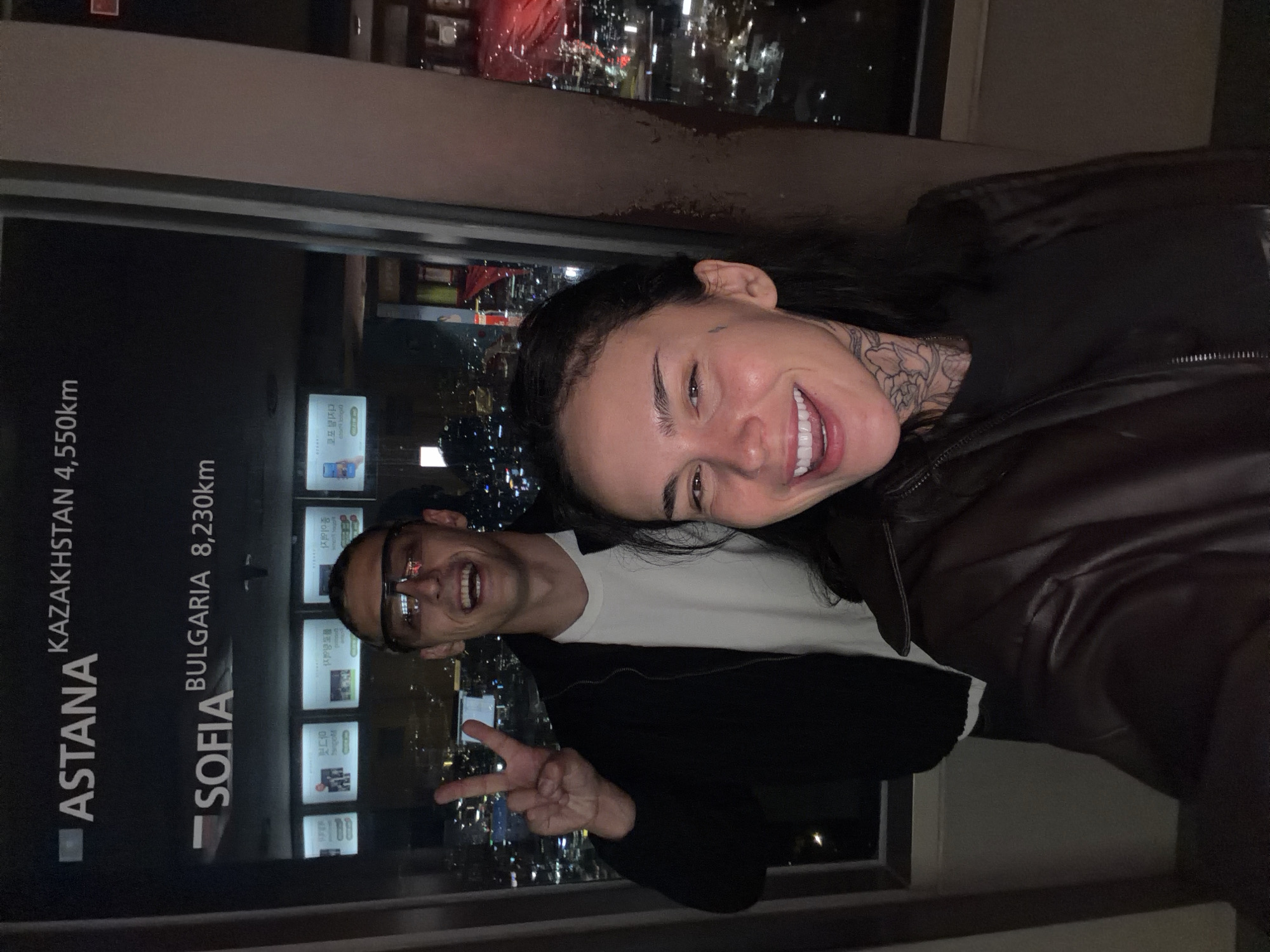 Two people are photographed in an indoor setting with a city visible through a glass panel in the background. The woman, positioned towards the right of the frame, is fair-skinned with dark hair and neck tattoos, wearing a dark leather jacket. She is smiling broadly. Next to her, a man with glasses gives a peace sign.
To the left is a sign reading "ASTANA KAZAKHSTAN 4,550km" and "SOFIA BULGARIA 8,230km." The background reveals a nighttime city landscape with numerous lights, characteristic of urban environments, suggesting the city is Seoul, South Korea, based on the information plaques in the background.