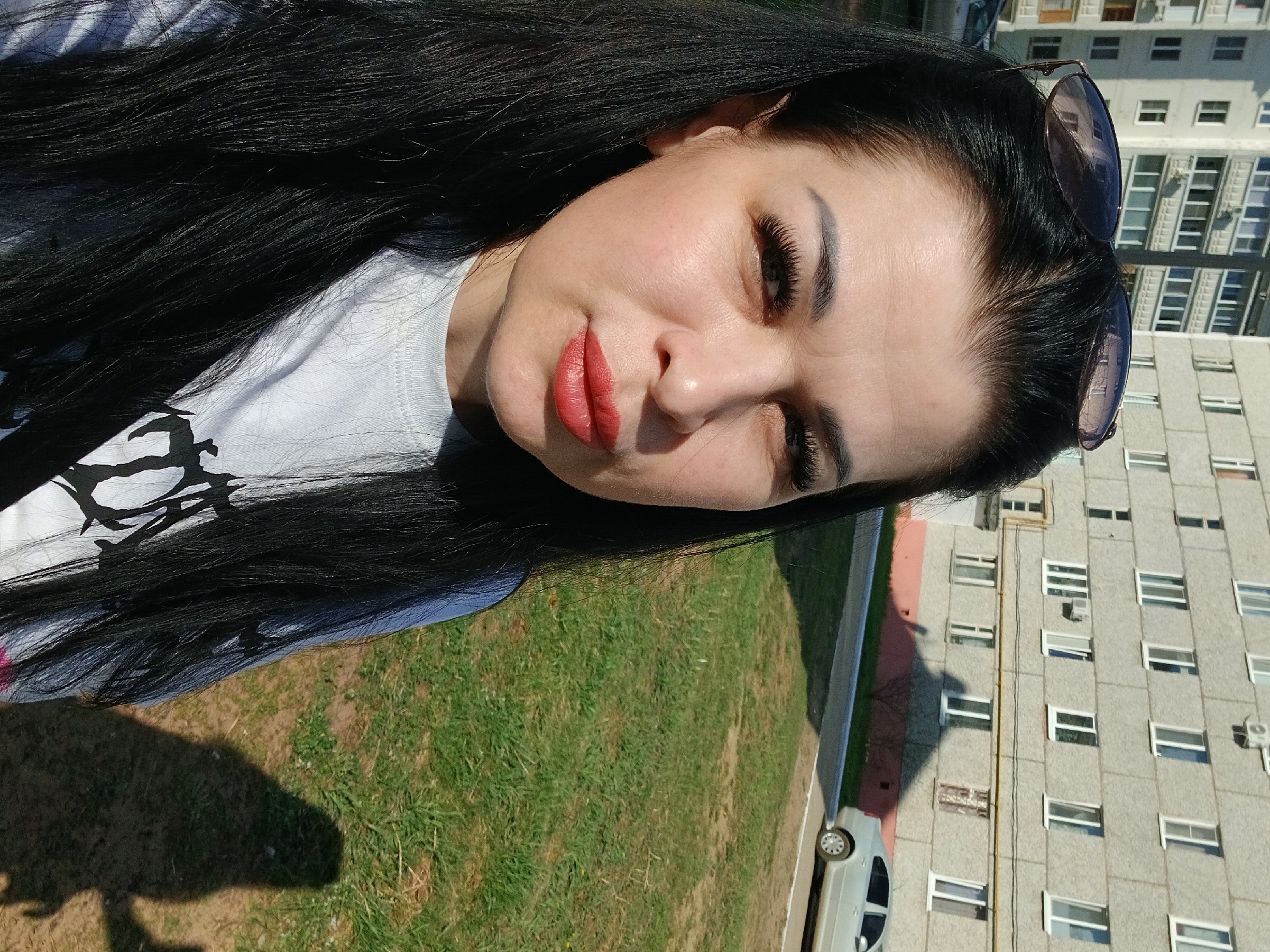 A woman with long black hair and red lipstick is the primary subject of the image. Sunglasses are perched on her head. She's wearing a white t-shirt with a black design. The setting appears to be outdoors in Noviy, Russia, as indicated by the presence of green grass, a parked car, and a multi-story apartment building in the background. The building has numerous windows and an exterior wall with a gray hue.