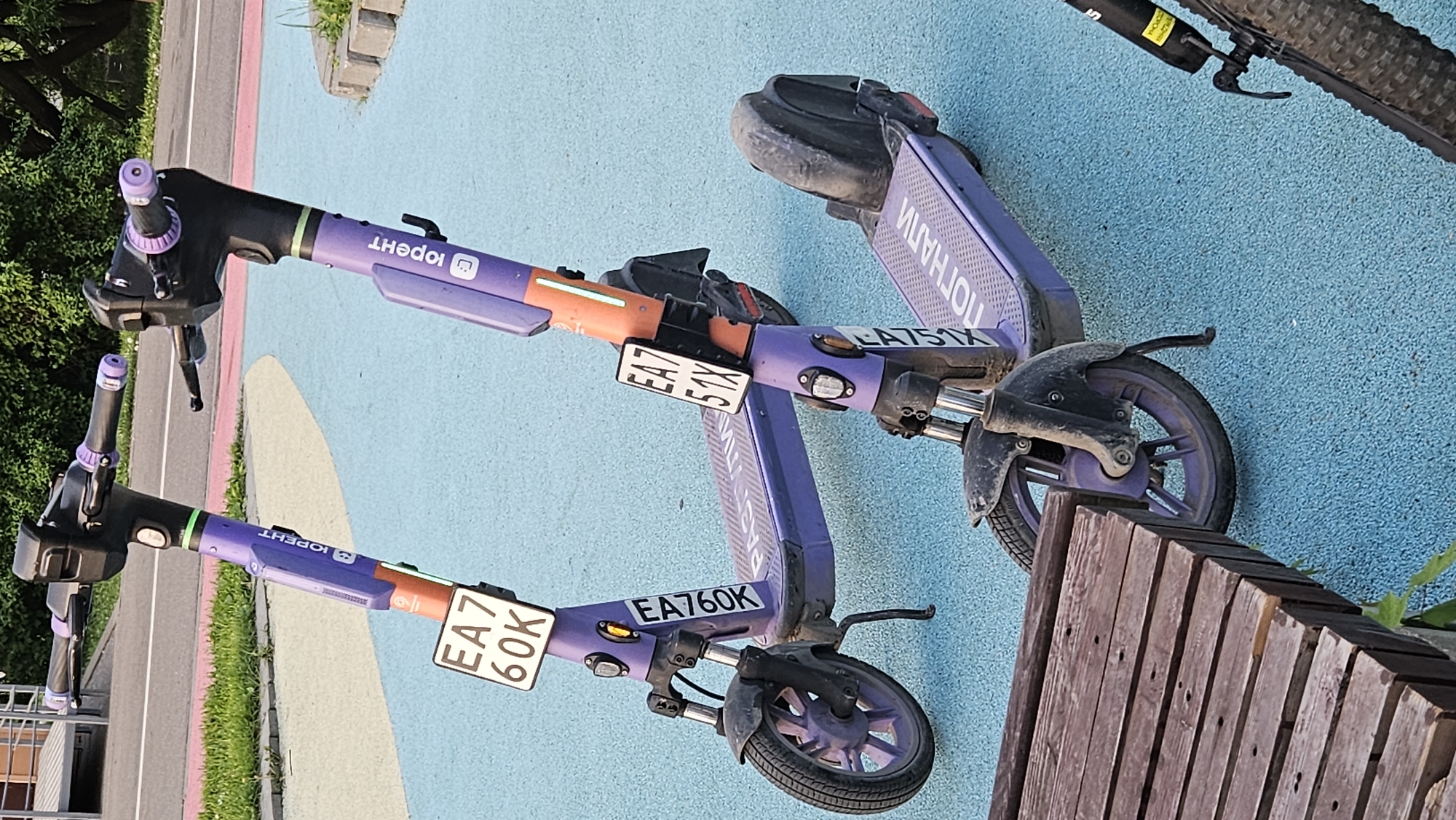 Here's a detailed description of the image:
**Location:** The image appears to be taken on a paved area with distinct sections of color: light blue, beige, pink and grey. The exact city or country is unknown.
**Objects:**
* Three electric scooters, identifiable by the logo "IOPENT" on their frames. The scooters are mostly purple and black. Two are fully visible and one is cut off at the top of the picture.
* Each scooter has an attached license plate. Visible license plates read "EA7 60K", "EA7 51X", and "EA751X."
* A wooden bench is partially visible on the right side of the image, obscuring the wheels of one scooter.
* There is a portion of a bicycle tire visible at the top right corner of the image.
* Vegetation (trees, grass) is on the left side of the image.
* Some concrete tiles are also visible in the background.
**Environment:**
* The scene is outdoors, likely a designated scooter parking or staging area.
* The lighting indicates it is daytime.
**Inferences:**
* The scooters are likely part of a rental service.
* The scene is in a public space, possibly a park or recreational area.
* The scooter plates suggests the scooter's location might be from an European Union country.