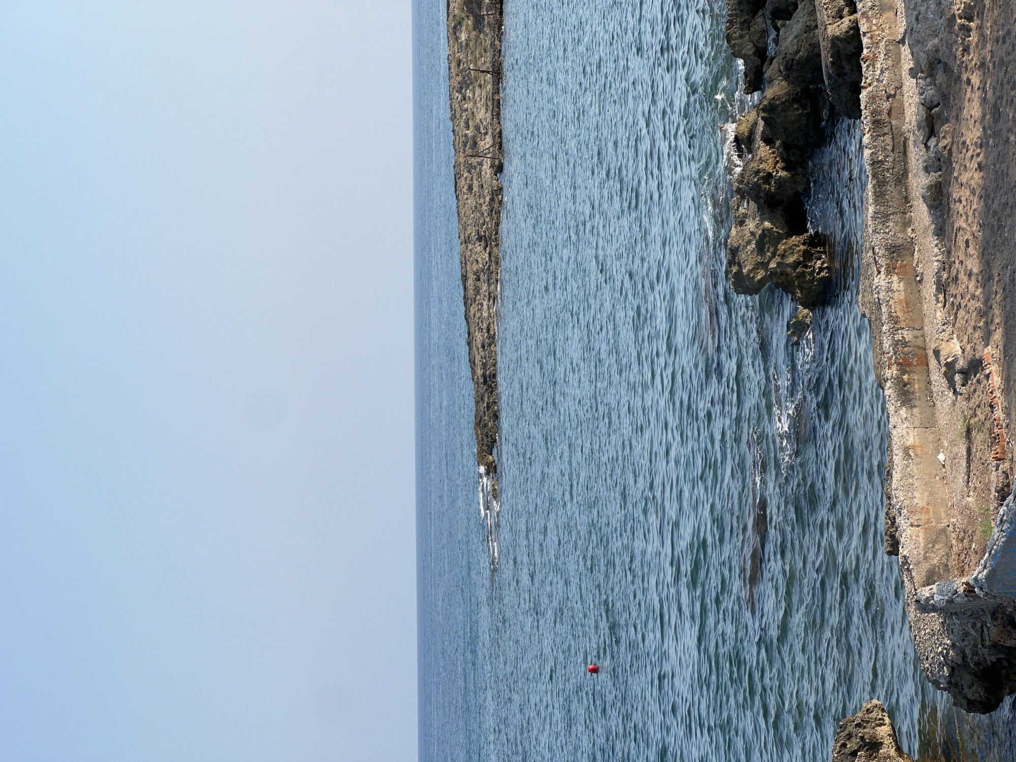 The image depicts a coastal scene with the clear blue sky meeting the calm blue sea. In the foreground, there are rough, sandy shores with scattered rocks and remnants of concrete structures, suggesting a natural or undeveloped coastline. The sea extends to the horizon, where a rocky outcrop juts out from the water. A small red buoy floats in the water to the left of the frame. The overall impression is one of a tranquil, natural environment along the coast of Antalya, Turkey.