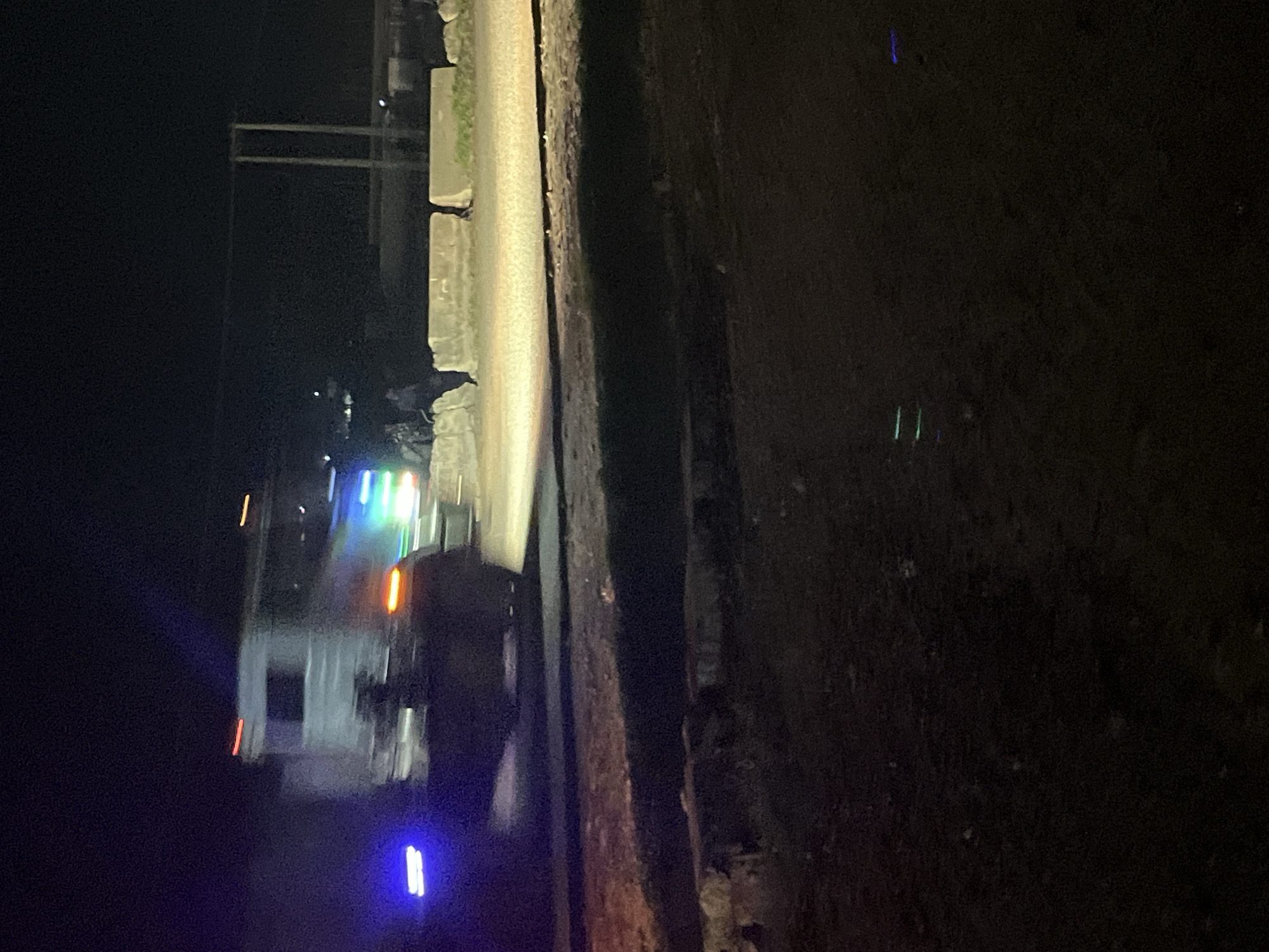 An unidentified truck, possibly a semi-trailer truck, is captured in motion at night on a paved road. The truck is illuminated by its own headlights and taillights, as well as vibrant blue and green lights mounted on its sides. The truck's chassis is white or light-colored, and it has a window on its side. The image also shows concrete barriers lining the side of the road, suggesting it might be a construction zone or a controlled area. Several indistinct figures are visible near the truck and the barriers, their identities obscured by the low light and motion blur. The environment appears to be an outdoor urban or semi-urban setting with a dark, unlit sky. The road surface is visible and appears to be asphalt.