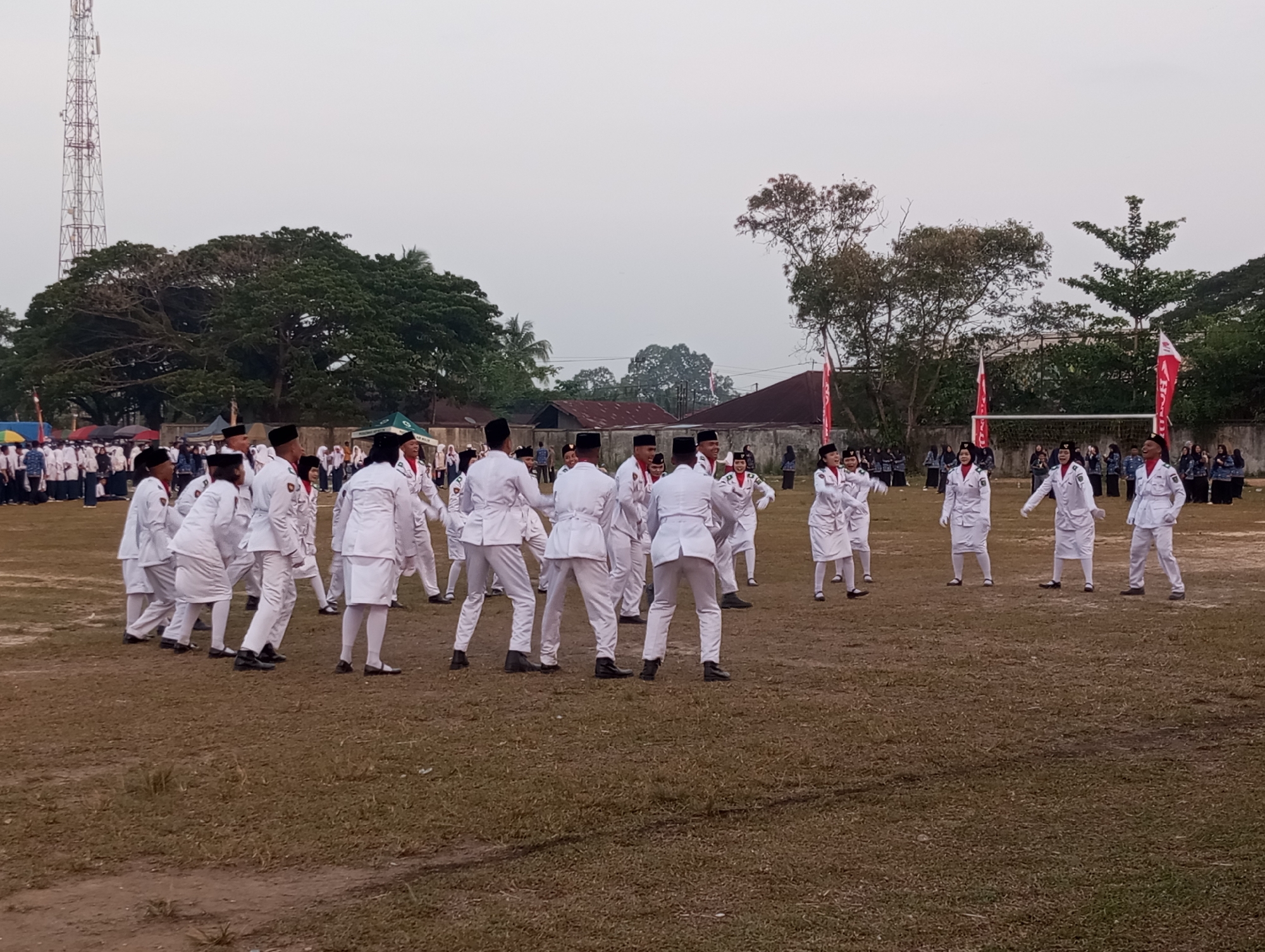 In Tanjung Gading, Indonesia, a group of people in white uniforms with red accents are performing a synchronized dance on a grassy field. The individuals, appearing to be students, are spread across the field in various formations, some with arms outstretched or bent at the elbows. They are wearing white long-sleeved jackets with matching pants or skirts, and dark shoes. Some have black hats. Red flags on poles are visible in the background, along with trees, buildings, and a tall communication tower. The sky is overcast.