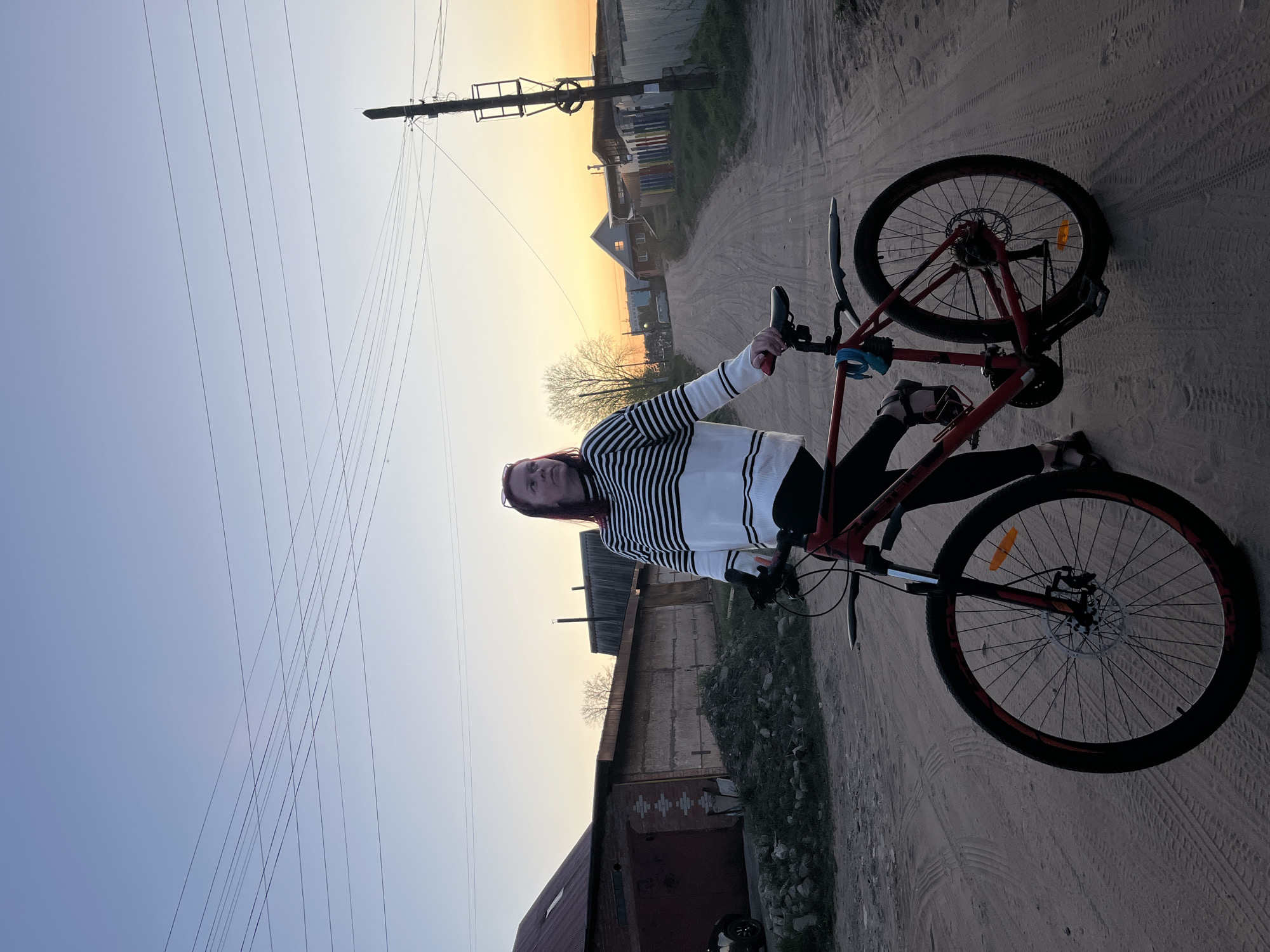 The image shows a person standing beside a red mountain bicycle on a dirt road. The person is wearing a black and white striped shirt and black pants. The dirt road appears to be located in a rural or suburban area in Chaikovsky, Russia, as evidenced by the presence of utility poles and buildings in the background. The sky is transitioning from blue to orange, suggesting it is either sunrise or sunset. The bicycle is stationary next to the person.