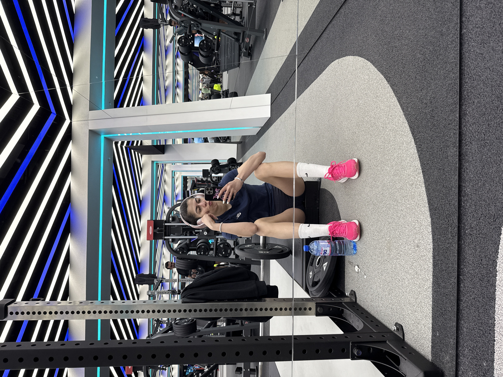 A gym setting in Sofia, Bulgaria shows a woman seated on the floor, facing a large mirror. She is wearing a dark blue athletic shirt, shorts, white socks, and bright pink sneakers. She holds a smartphone in her right hand, presumably taking a selfie. A water bottle and weights surround her on the floor. Behind the woman is a weight rack with black weights, and beyond that, various gym equipment. The walls and ceilings are decorated with geometric patterns of black, white, and blue lines, giving the gym a modern aesthetic.