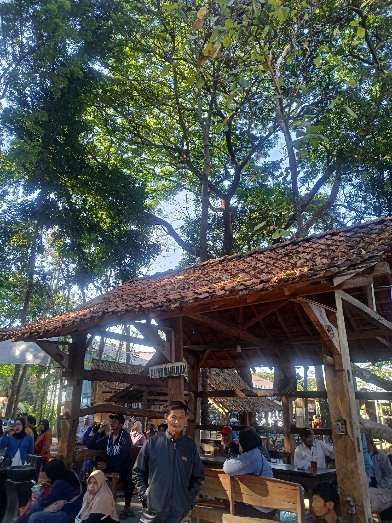The image depicts a busy open-air market scene in Karungan, Indonesia. In the foreground, a man in a dark jacket stands facing the camera. Behind him, a rustic wooden pavilion with a tiled roof provides shelter. Numerous people are seated at wooden tables and benches, some consuming drinks, while others are standing or walking around. A sign above reads "PASAR BAHULAK," indicating the name of the market. The background is filled with lush green trees, suggesting a natural, possibly rural or park-like, setting. The overall atmosphere appears lively and social, with many individuals gathered at the market.