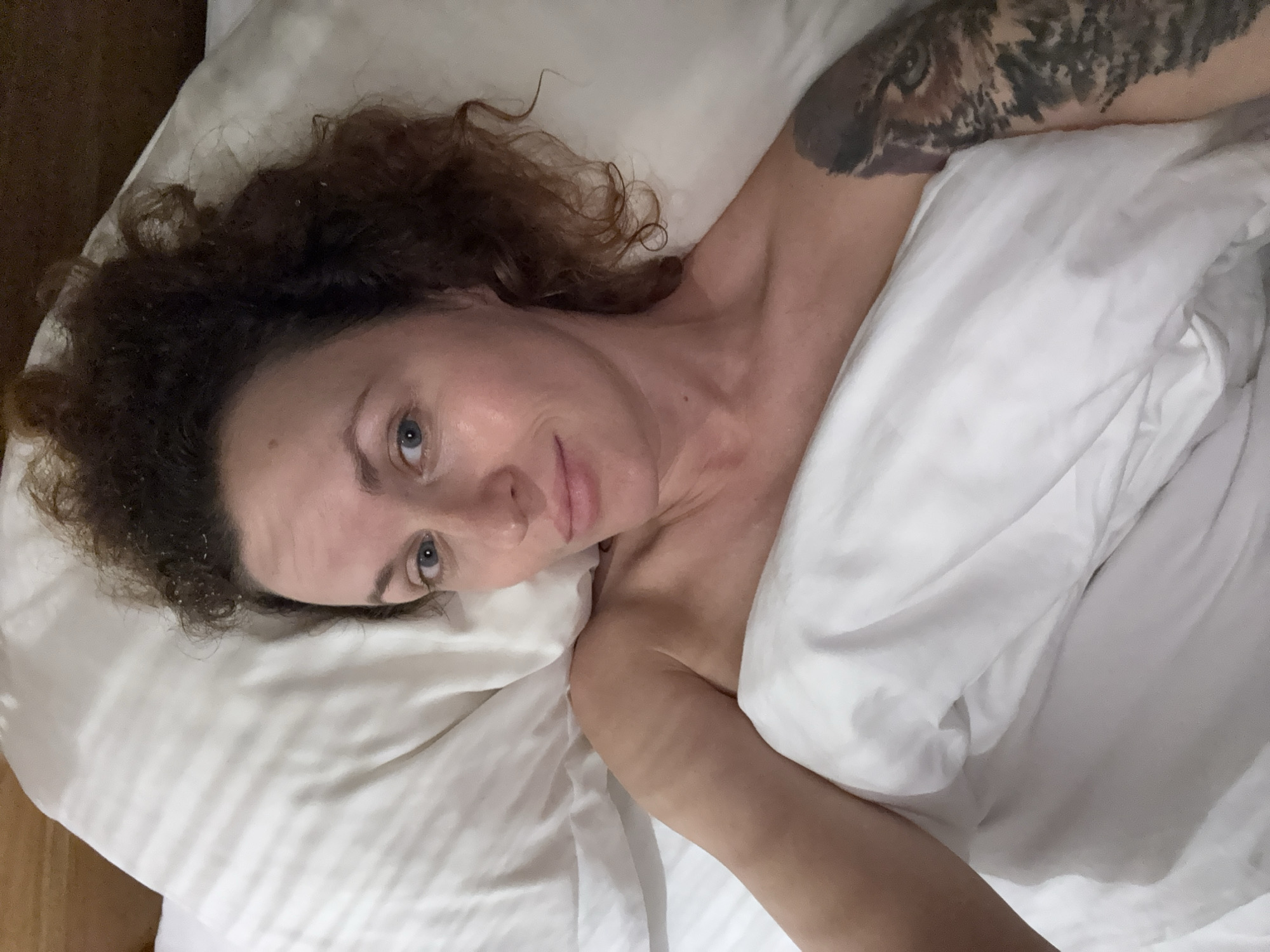 A woman with curly hair and a tattoo on her upper arm is lying in a bed with white linens. She is looking at the camera. The location is Mengwi, Indonesia.