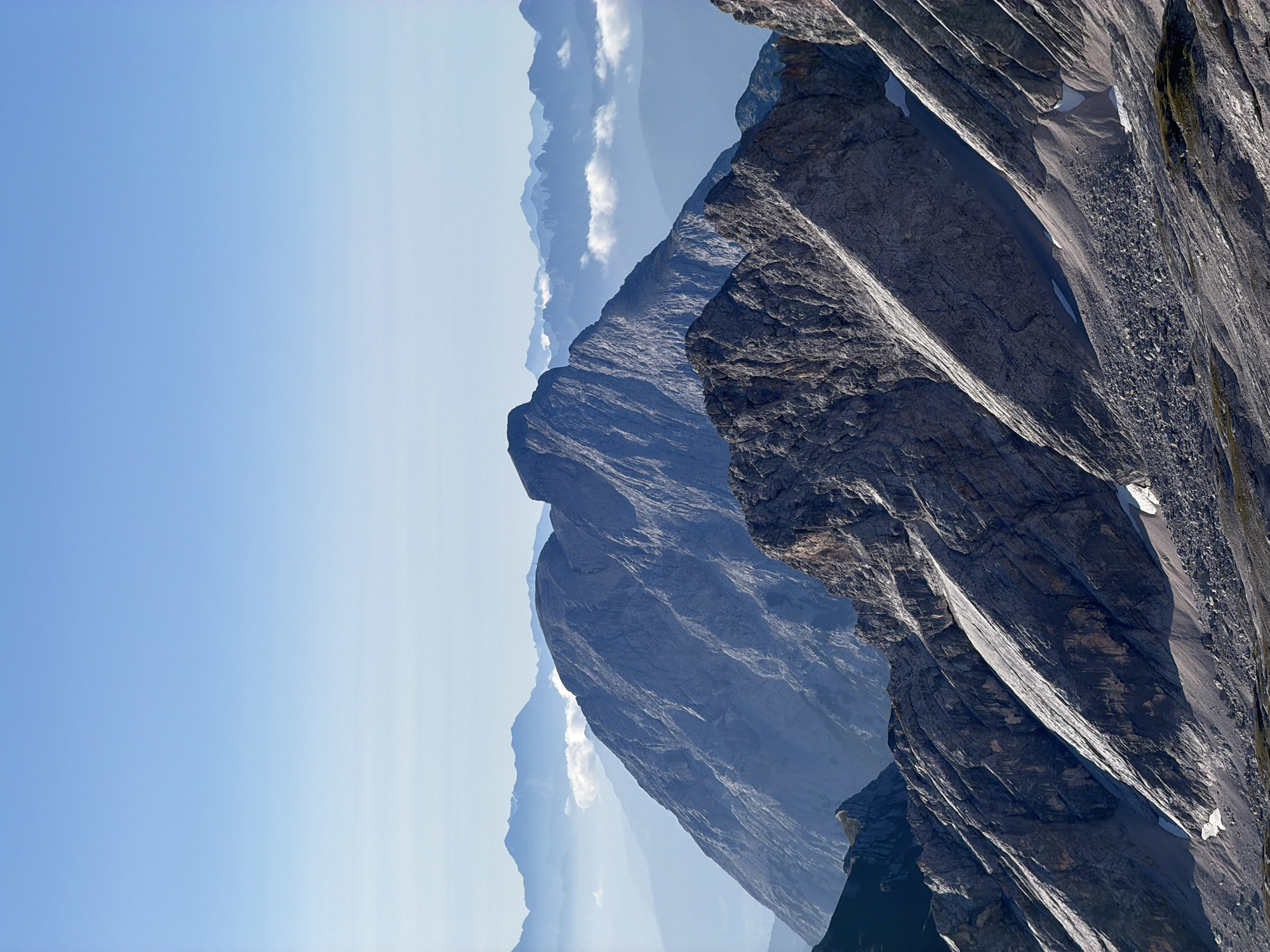 This is a vertical, eye-level, medium shot of a mountainous landscape. The foreground features rugged, rocky slopes with patches of snow and scree. Diagonal lines of rock formations dominate the lower half of the image, leading the eye towards the midground.
In the midground, a prominent, sharp-peaked mountain rises, its steep, textured slopes illuminated by the sun. Beyond this, a series of progressively softer, blue-hued mountain ranges recede into the distance, creating a sense of depth. Wisps of clouds are scattered between the midground and background mountains, adding to the atmospheric perspective.
The sky above is a clear, gradient blue, transitioning from a lighter shade near the horizon to a deeper hue at the top of the frame. There are no visible people, animals, or man-made structures in the image, focusing entirely on the natural mountain environment. The scene suggests a high-altitude location, likely in the Bavarian Alps. While the image does not explicitly depict Garmisch-Partenkirchen, the described landscape is characteristic of the mountainous region surrounding the city.