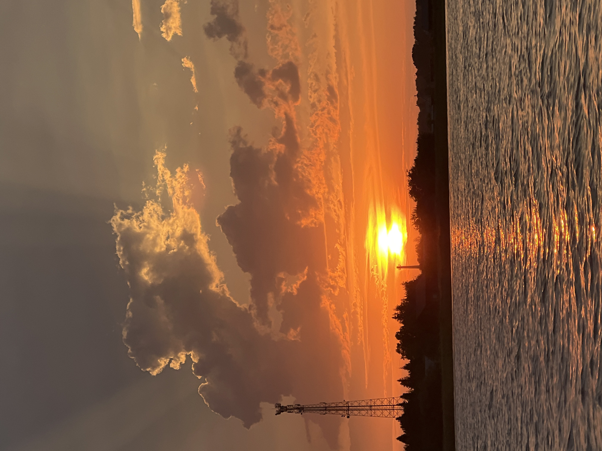 A cell tower stands tall to the left of a body of water, silhouetted against a vibrant sunset over Novopichugovo, Russia. The sky is filled with dramatic clouds illuminated by the setting sun, casting a warm orange glow across the scene. The sun itself is a bright orb near the horizon, with rays of light fanning out behind it. Trees and buildings are visible along the shoreline, their forms darkened by the fading light. The water reflects the colors of the sunset, creating a shimmering effect on its surface.