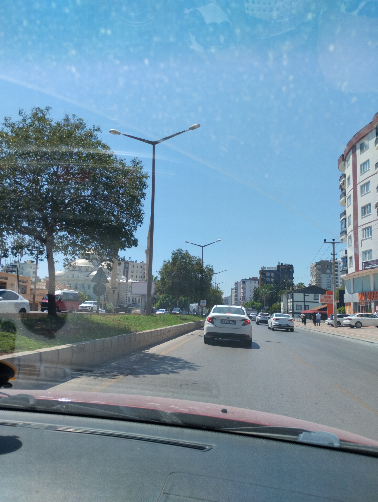 The image depicts a street scene in Mezitli, Turkey, on a bright, sunny day. The perspective is from inside a vehicle, looking forward through the windshield. The street is paved and appears to be a multi-lane road with traffic moving in the same direction as the viewer's vehicle.
Several cars are visible on the road. Closest to the viewer, a white car with license plate "01 AZV 219" is prominent in the right lane. Further ahead, other white cars and a silver car are driving. Pedestrians can be seen walking on the sidewalk on the right side of the street.
Buildings line the right side of the road, ranging in height and architectural style. Some appear to be modern apartment buildings, while others are commercial establishments. One building prominently features the signage "MM MIGROS".
On the left side of the road, a wide median strip is covered with grass and trees. A large, mature tree with dense foliage is a significant element in the mid-ground on the left. Beyond the median, there is a view of a building with a dome, possibly a mosque, with minarets visible as well.
Streetlights with multiple lamps are positioned along the road. The sky is clear and blue with some reflections and specks visible on the windshield. The overall impression is of a typical urban environment in a coastal city during daylight hours.