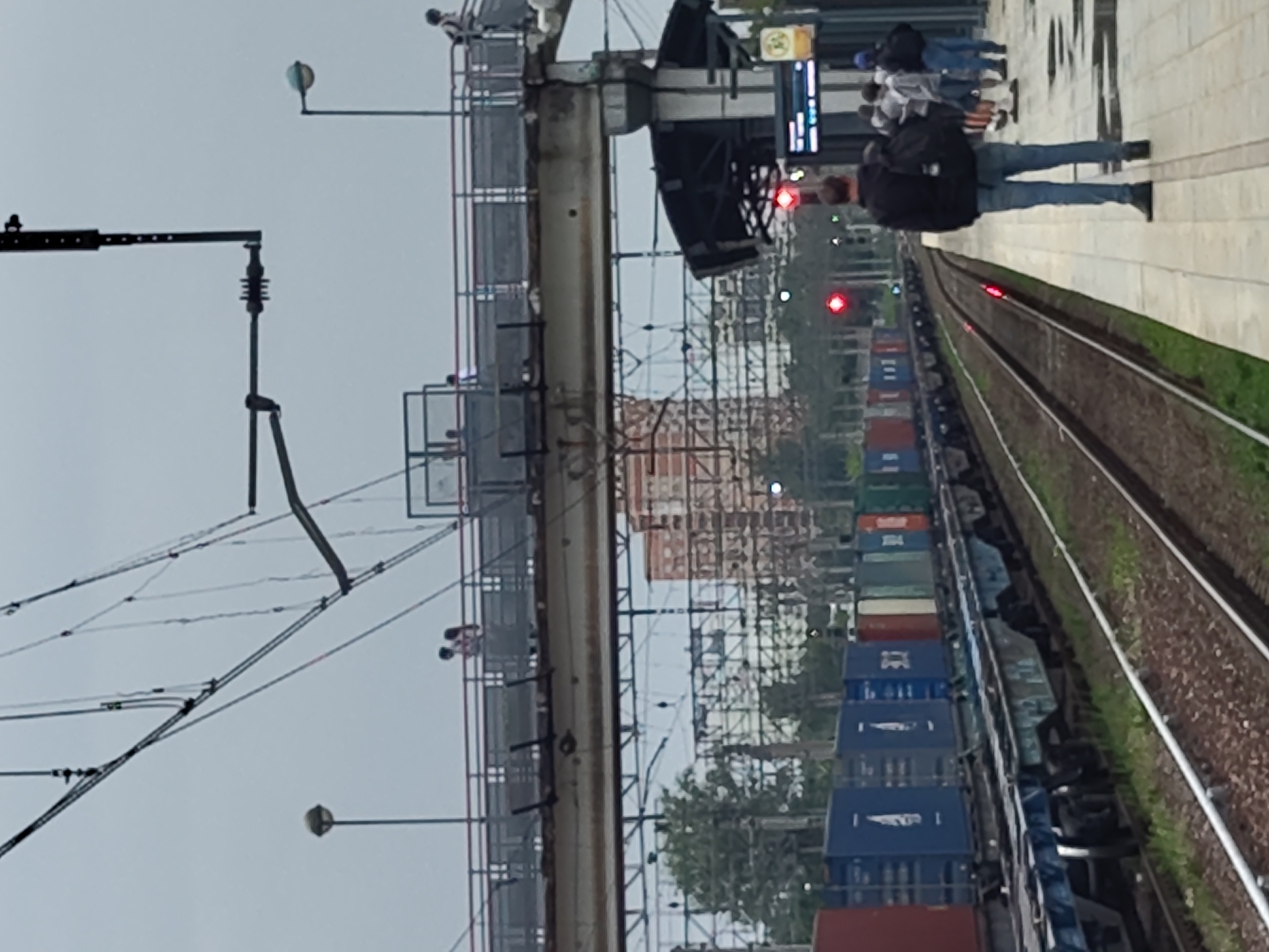 The image depicts a train station platform in Ramenskoye, Russia, under an overcast sky. A freight train, laden with shipping containers of various colors including blue, red, and green, is on the tracks to the left. Overhead, a complex network of electrical wires for the train system is visible. A pedestrian bridge spans across the tracks above the train. On the bridge, a few silhouetted figures can be seen. On the platform in the foreground, a man wearing a black hooded jacket and blue jeans stands facing the tracks, with a backpack on his shoulders. Several other individuals are visible further down the platform, some standing and some walking. A signal light, showing red, is illuminated near the tracks. A station information display board is mounted on a pole to the right. The platform itself is paved with light-colored tiles, and some areas appear wet, suggesting recent rain. The background shows trees and a building, likely in Ramenskoye.