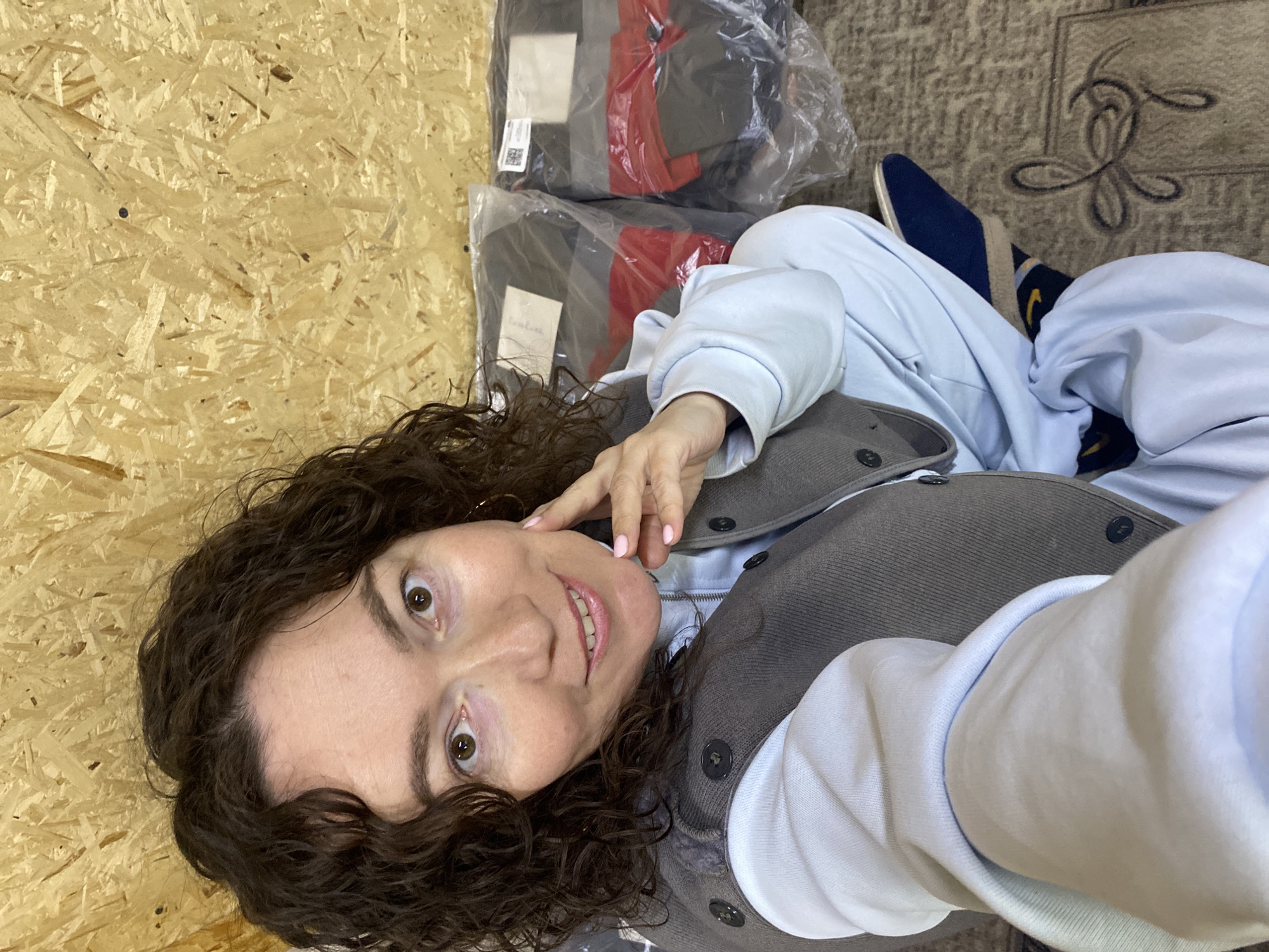 A woman with curly brown hair is seen from a low angle, likely a selfie, in what appears to be an indoor setting. She is wearing a light blue tracksuit and a grey waistcoat with buttons. The background includes a wooden panel, possibly a wall, on the left, and a carpet with a floral pattern on the right. Two wrapped packages are visible in the upper background, containing items with red and grey colors. The woman is touching her face with her right hand and smiling. Blue slip-on shoes are visible in the upper right. The location is Ol'khovskoye, Russia.