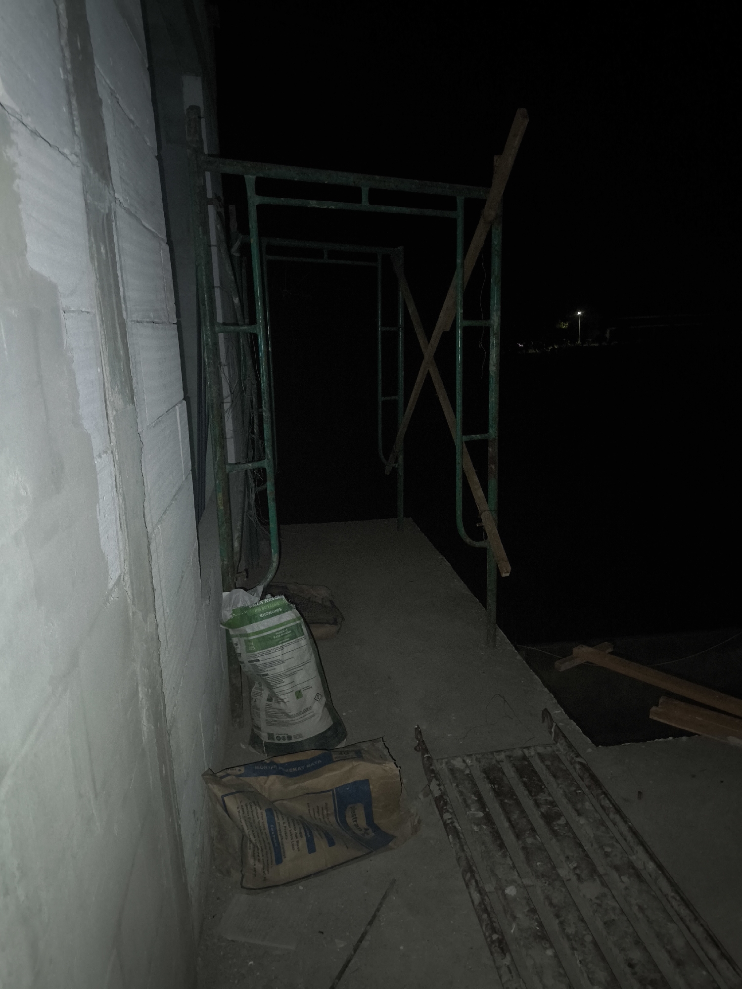 The image depicts a construction site at night. A white brick wall, partially covered in a light grey material, occupies the left side of the frame. Adjacent to the wall is a green metal scaffolding structure with wooden planks forming diagonal supports. Two bags of building materials, one green and one brown, are placed on the concrete ground in front of the scaffolding. The ground is uneven and dusty. To the right of the scaffolding, a section of the ground appears to be a metal grating. In the background, a dark sky is visible with a distant streetlight and a few blurred figures.