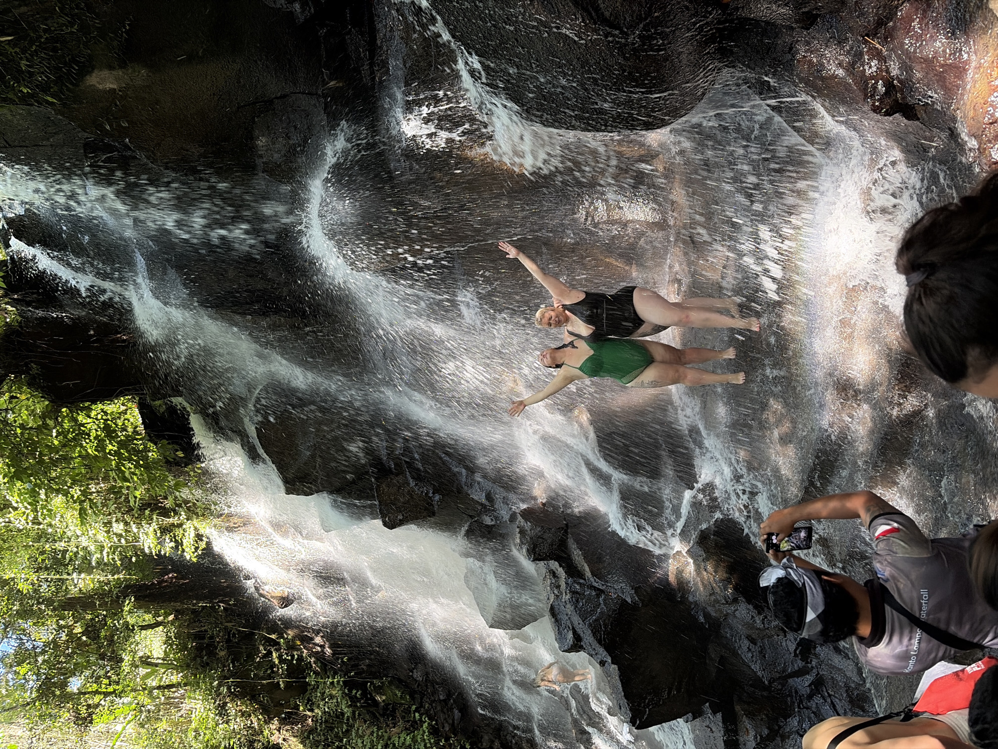This image depicts a waterfall located at Kanto Lampo Waterfall in Gianyar, Indonesia. Two women are standing under the waterfall, with their arms raised in celebration. The woman on the left is wearing a green one-piece swimsuit, and the woman on the right is wearing a black one-piece swimsuit. In the foreground, a person wearing a grey shirt with "Kanto Lampo Waterfall" printed on it is taking a picture of the waterfall with their phone. The environment is lush and tropical, with dense foliage surrounding the waterfall. The water cascades over dark, rocky formations, creating a dynamic and refreshing scene.