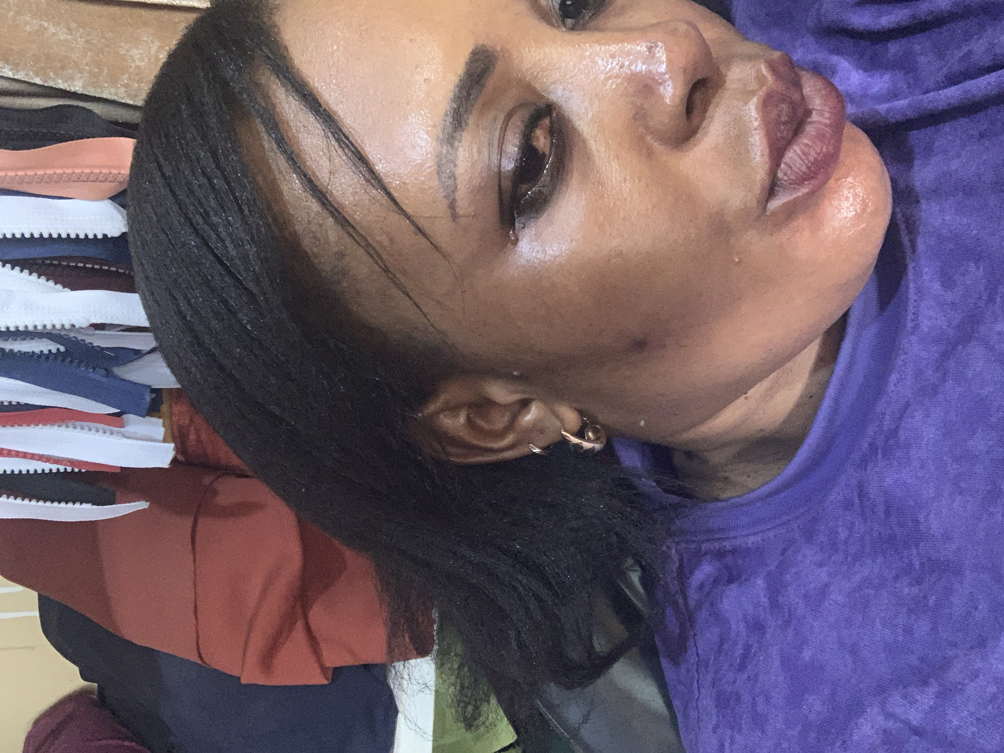 The image shows a close-up, head-and-shoulders shot of a Black woman, seemingly in a relaxed or resting position. She is wearing a purple t-shirt. Her hair is dark and straight, framing her face. A small gold hoop earring is visible in her left ear. The background is out of focus, but appears to be a stack of folded garments, including items with white, blue, and reddish-brown fabric.
The woman's face is turned away from the camera, with her gaze directed downwards and to the left. Her skin appears to be dewy, possibly from sweat or skincare. The lighting suggests an indoor setting, possibly during the daytime. No specific activity is clearly depicted, but the pose could suggest resting or looking at something out of frame.
Notable details include the texture of her skin, the sheen on her face, and the fabric patterns in the background. No text is visible in the image. The location context is Sabon Tasha, Nigeria.